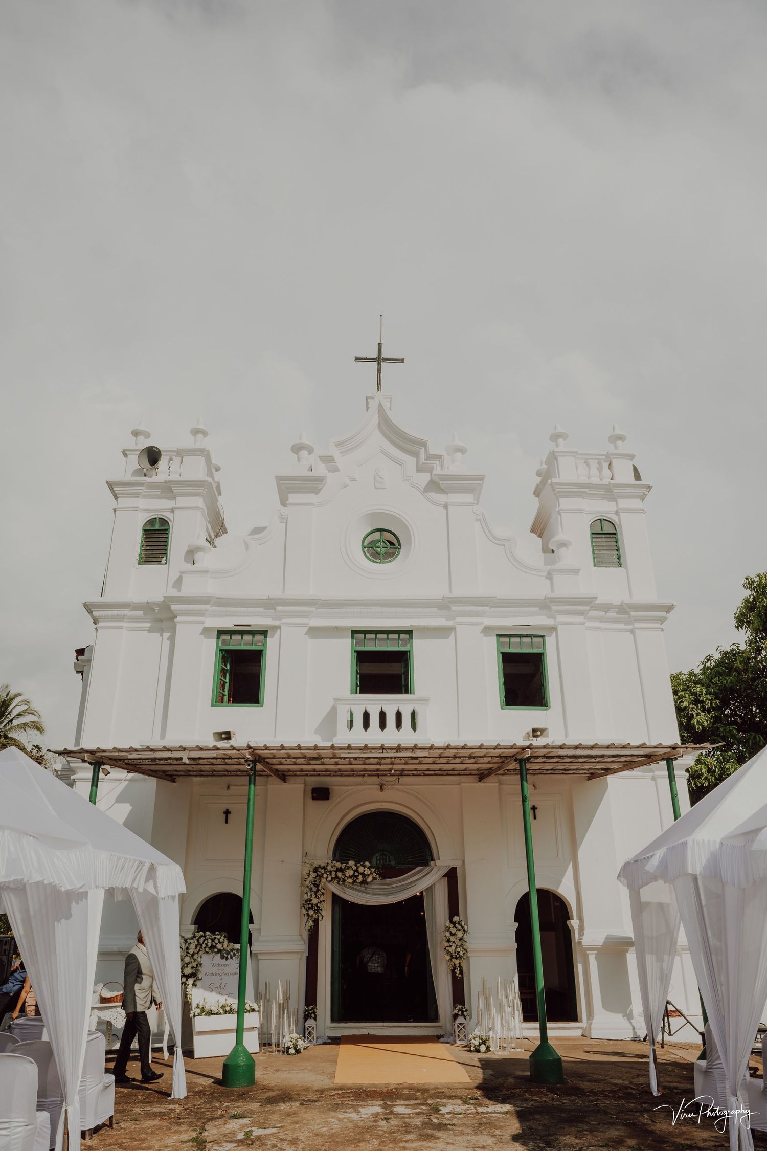 White colonial Portuguese church with three towers and cross stands behind temporary white tents against cloudy sky.