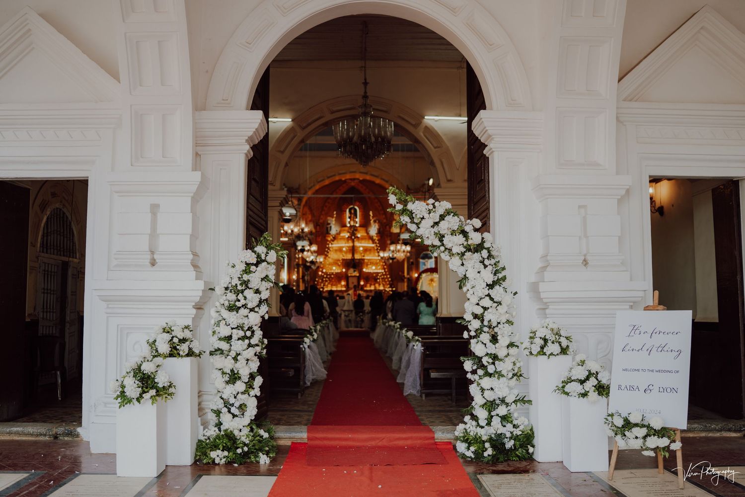 White floral wedding arch decorates the entrance to a church with a red carpet leading inside through arched doorways.