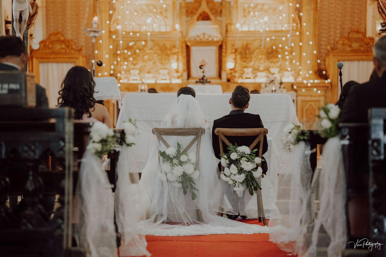 Couple seated at church altar during wedding ceremony with white flowers and glowing lights decorating the backdrop.