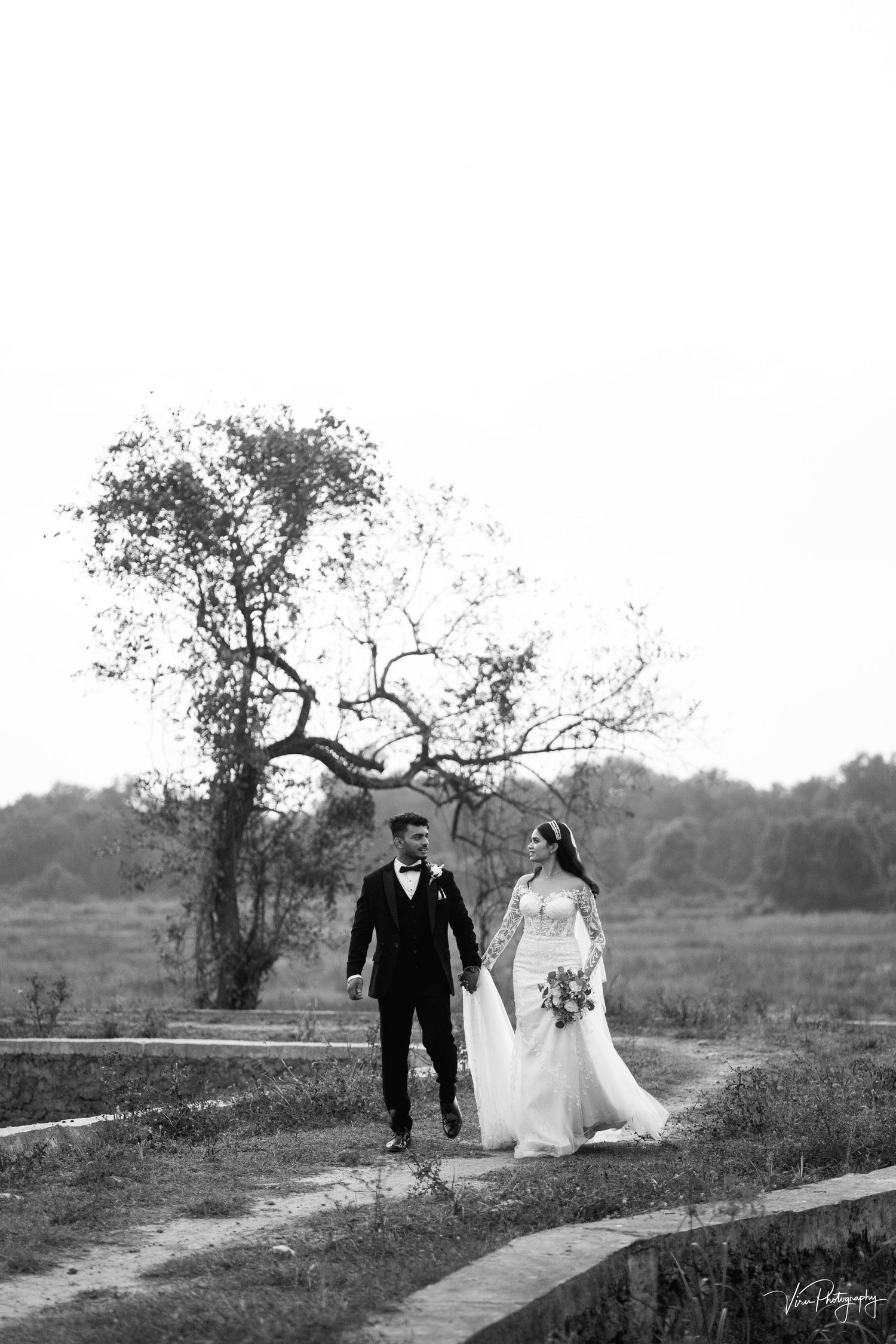 A romantic black and white wedding photo showing a couple walking along a winding path with a dramatic tree in the background.