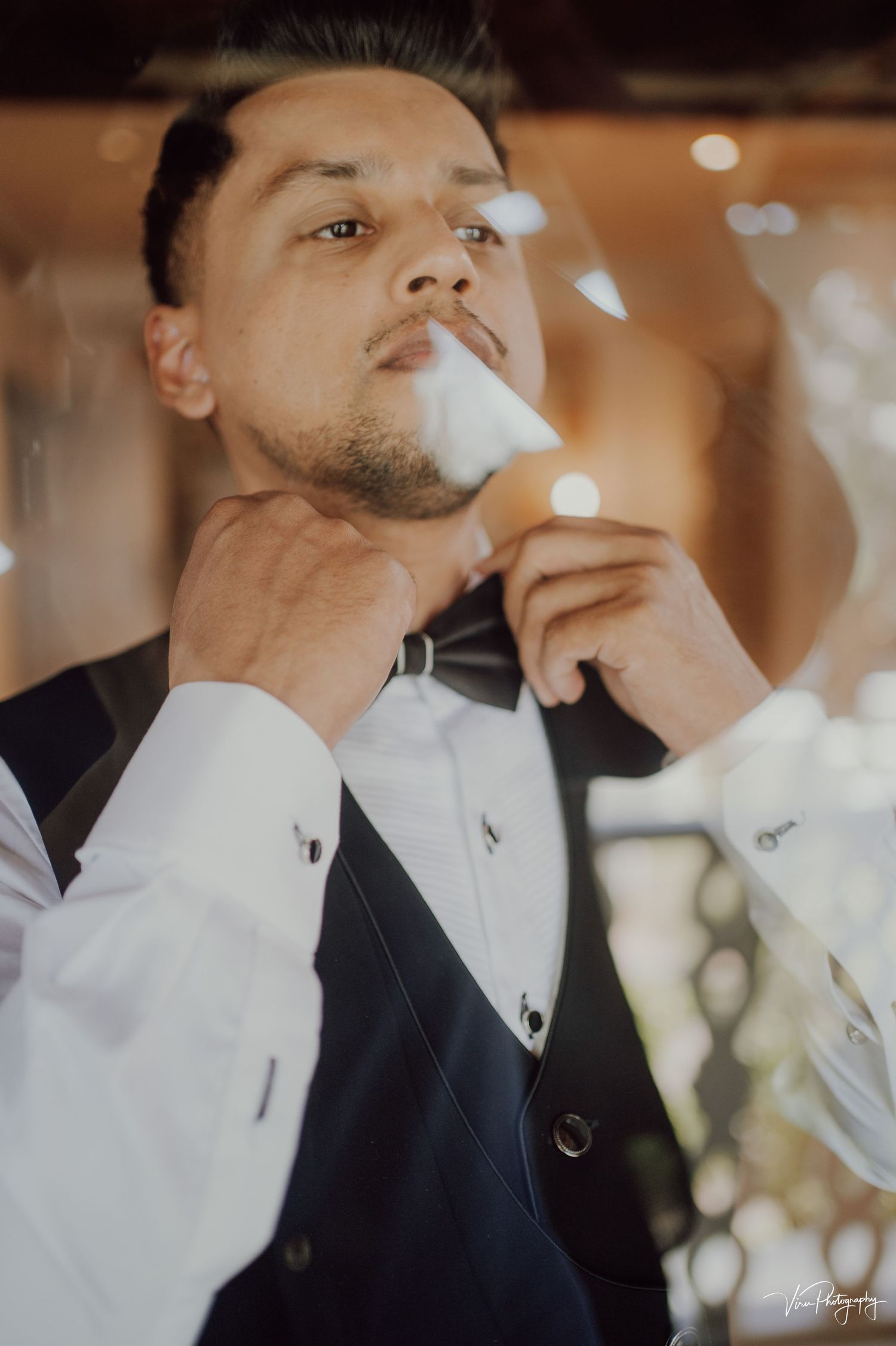 Well-dressed person in tuxedo and vest adjusts bow tie while exhaling smoke in warm ambient lighting.