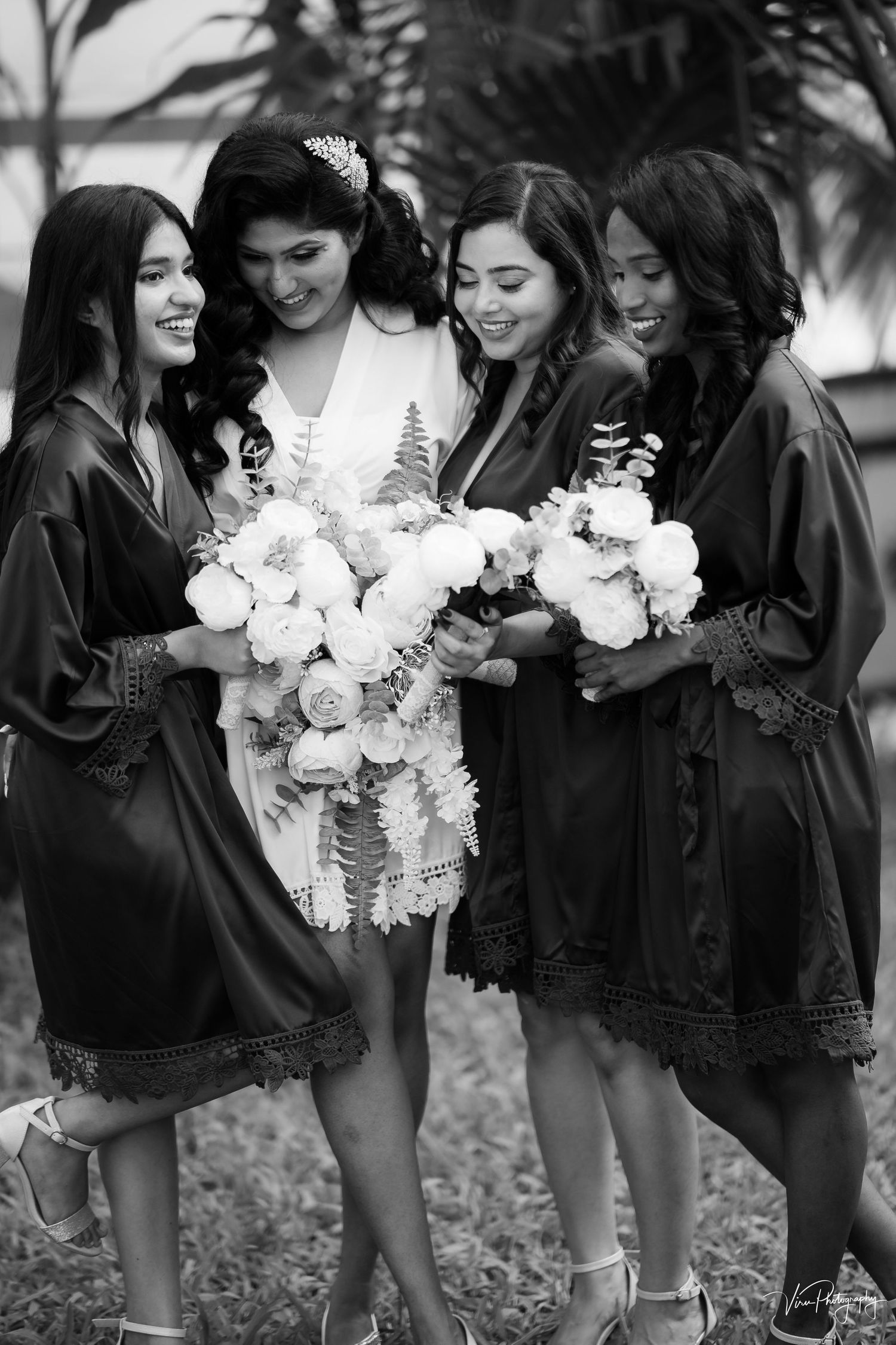 Black and white photo of bridesmaids in matching silk robes holding white floral bouquets during wedding preparations.