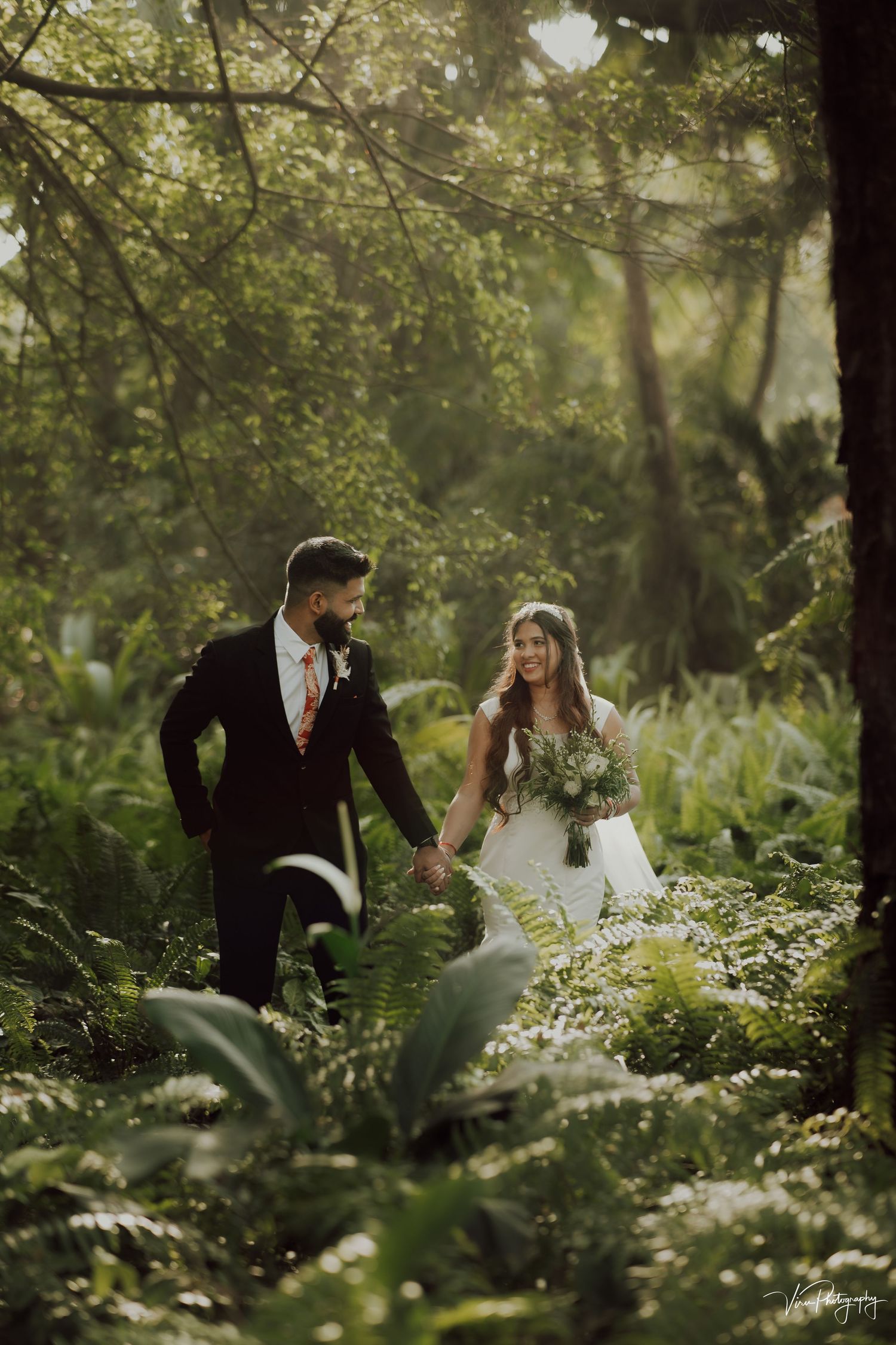 A romantic wedding photo of a couple walking hand-in-hand through a lush green forest setting in soft natural light.