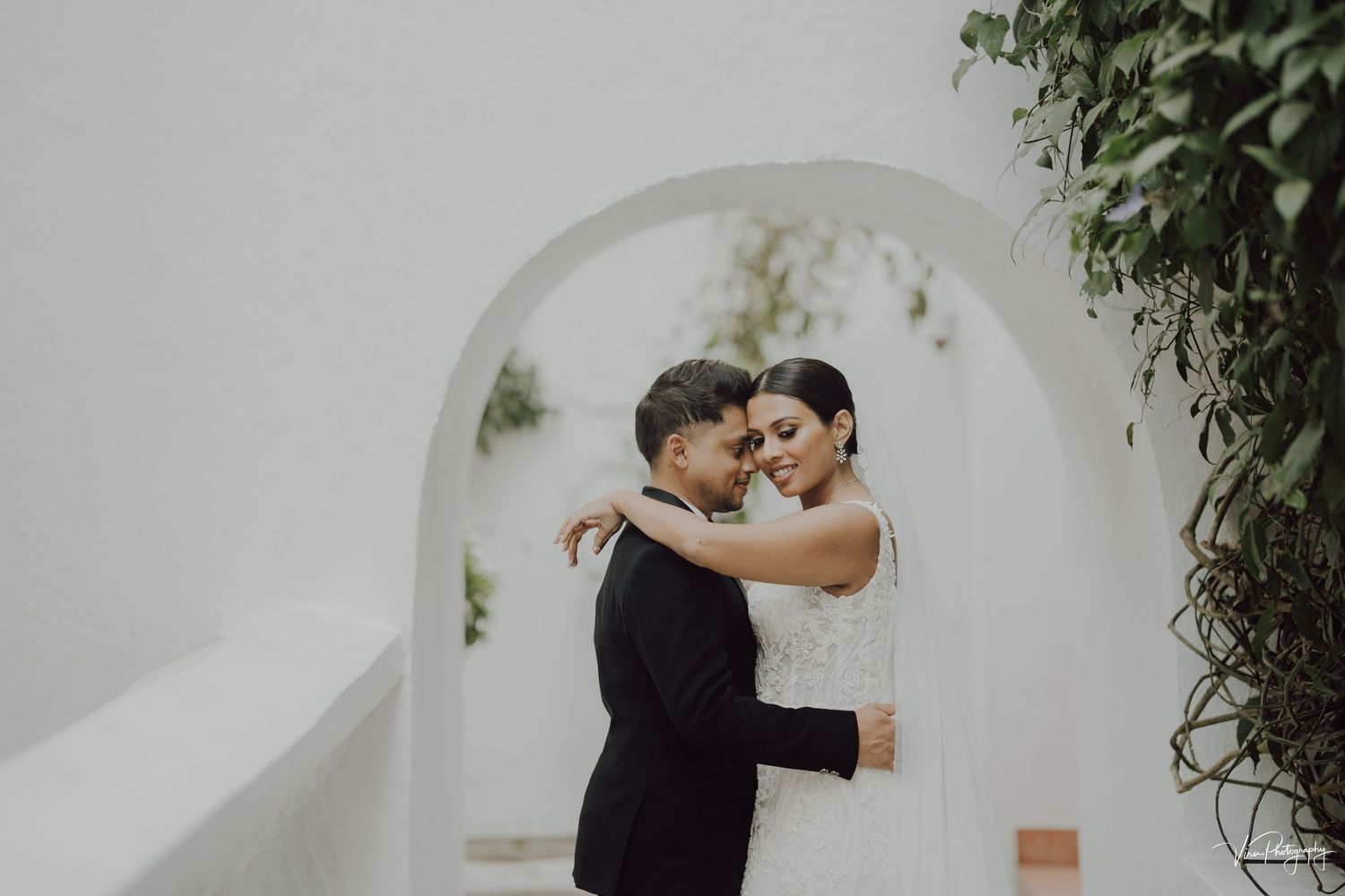 Romantic couple embraces under white archway decorated with greenery during wedding celebration.