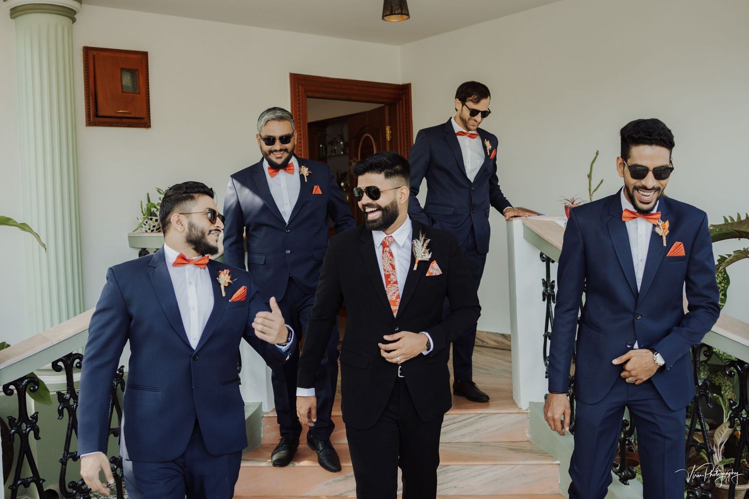 Groomsmen in coordinated navy blue suits and red accents share a candid moment at a wedding celebration.