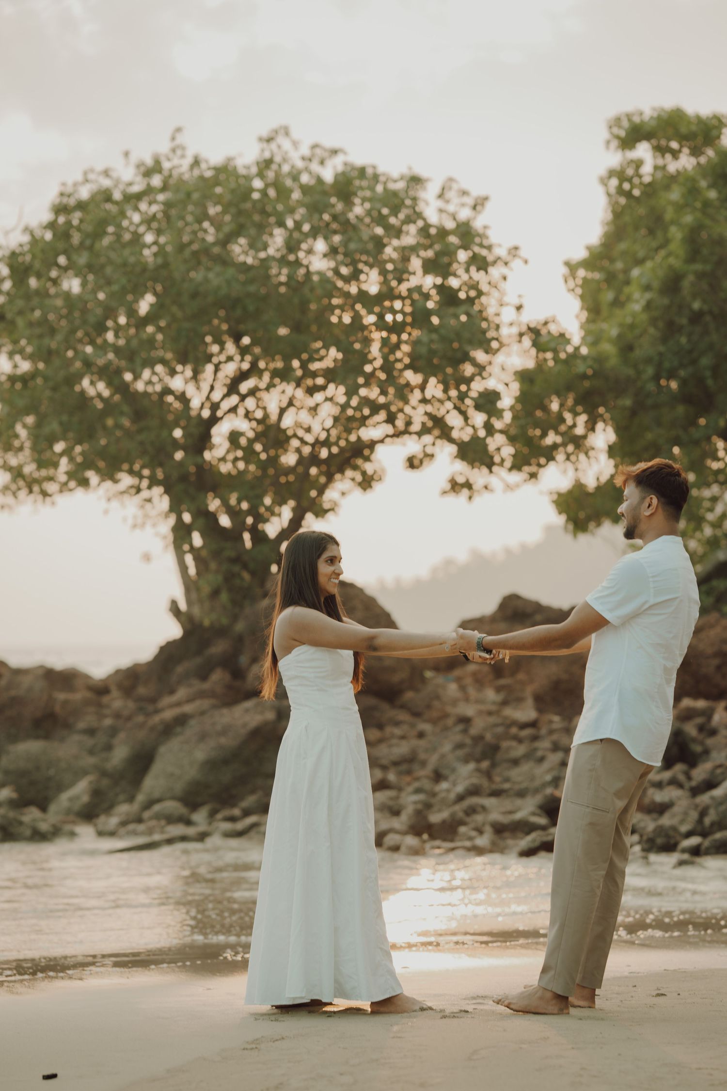 A couple holds hands and dances together on a sandy beach at sunset with trees and rocks behind them.