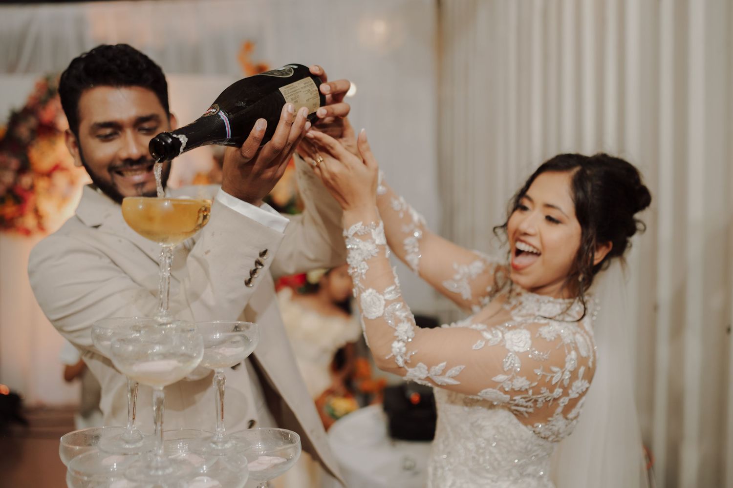 Bride and groom laugh together as they pour champagne over a tower of coupe glasses at their wedding celebration.