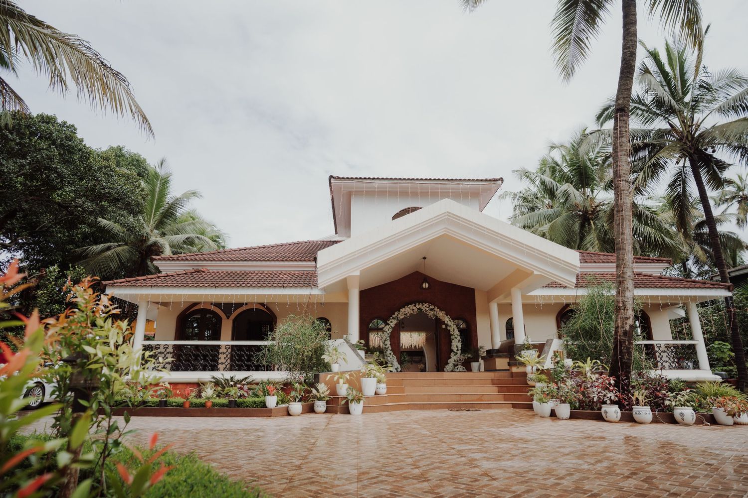 Charming Portuguese-style villa with red tiled roof, arched veranda, and potted plants set among tall palm trees on a cobblestone courtyard.