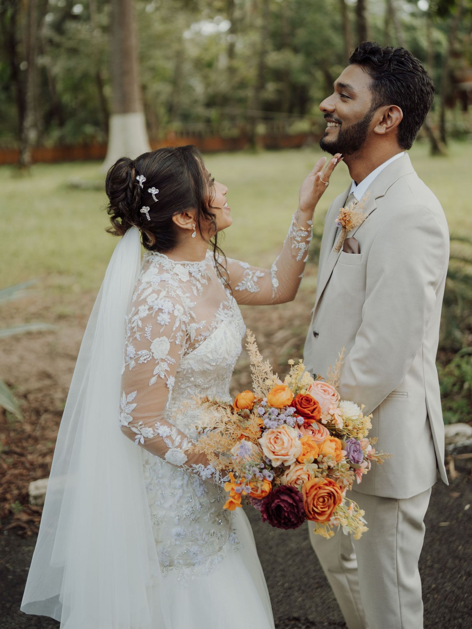 Smiling groom in a beige suit gently touches the cheek of his veiled bride holding an orange and pink floral bouquet outdoors.