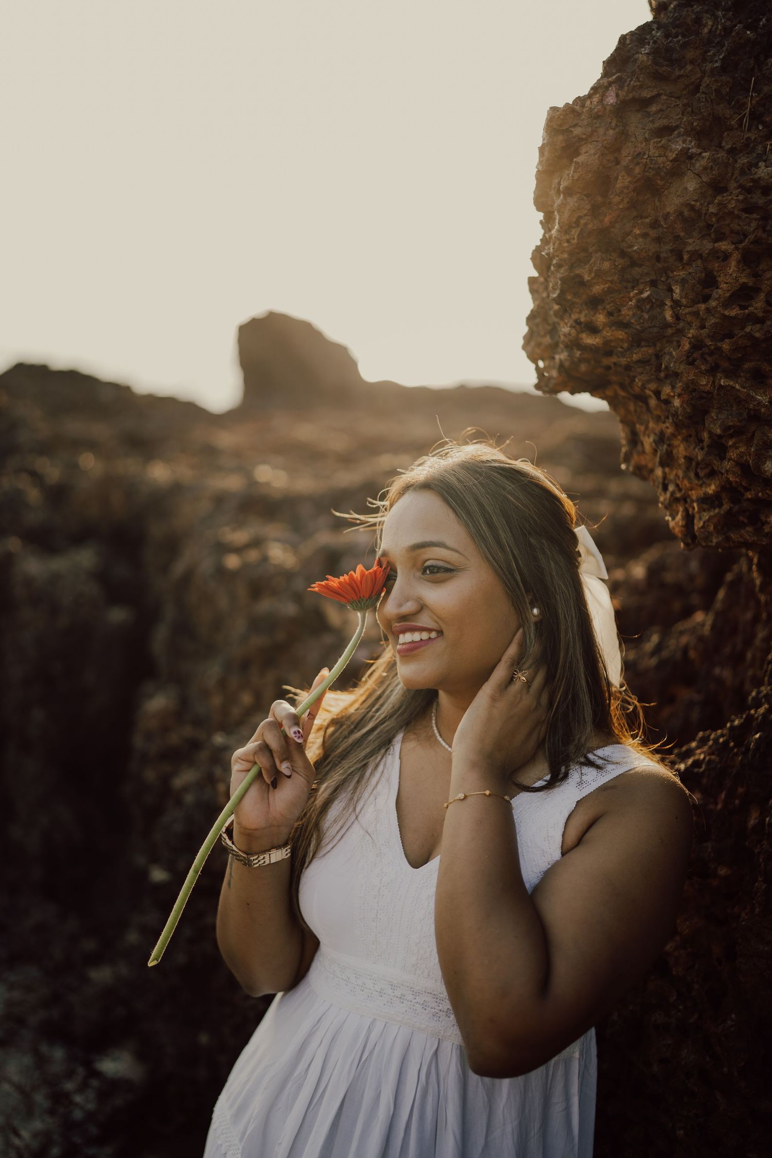 A person in a white dress poses against rocky cliffs at sunset holding a red flower in a dreamy photoshoot.