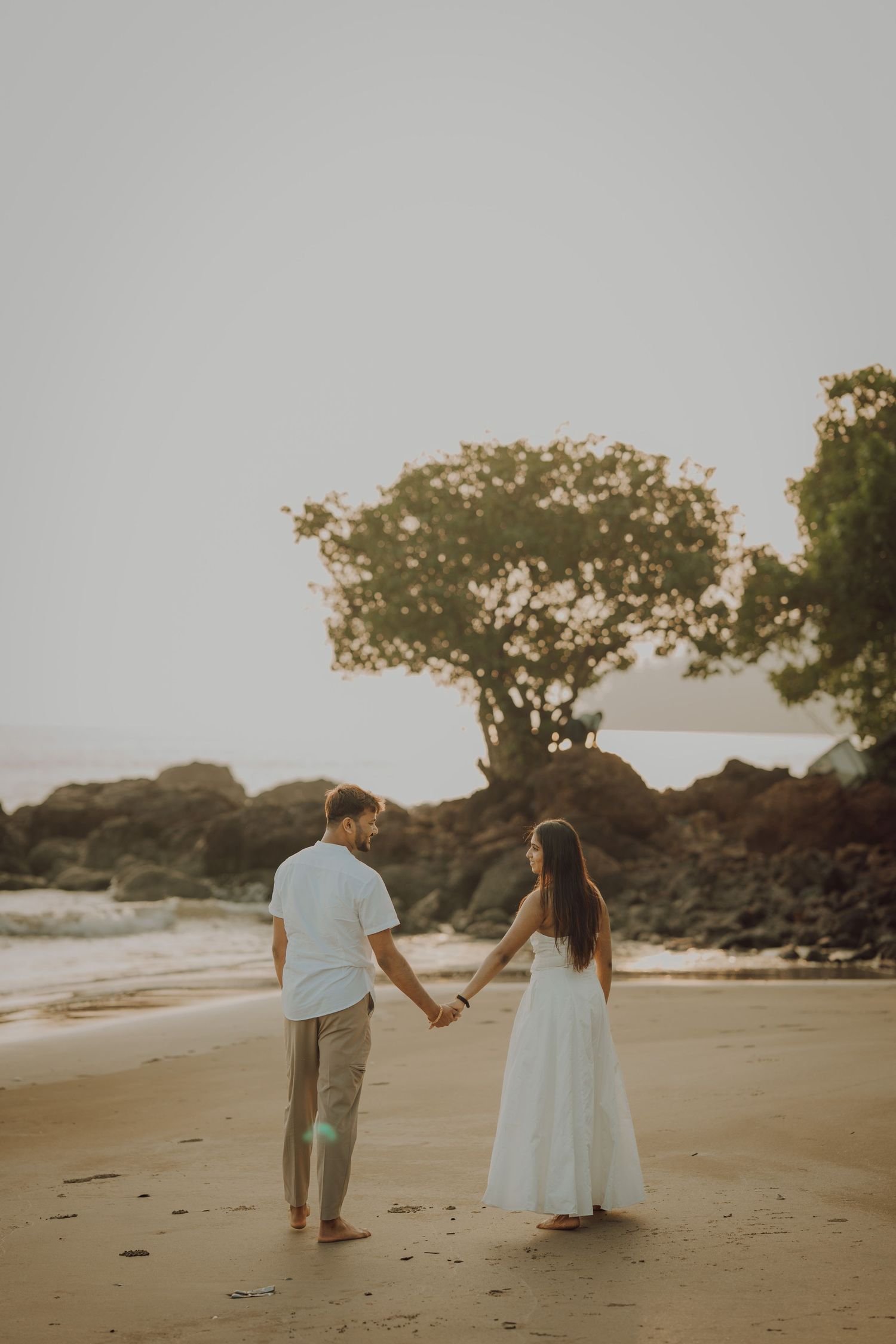A couple holds hands while walking along a serene beach at sunset with volcanic rocks and trees in the background.