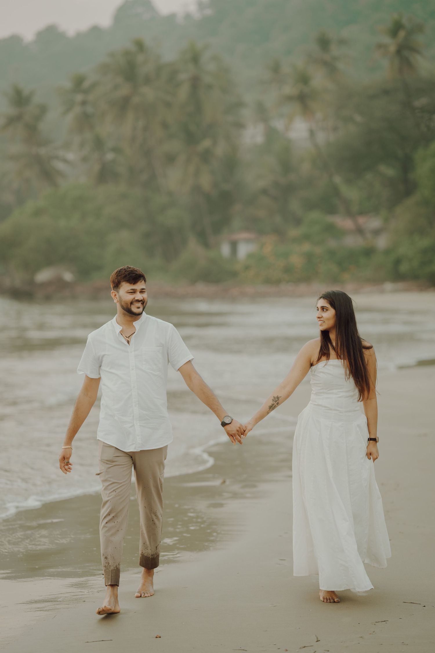 A couple walks hand in hand along a tropical beach with palm trees in the background during golden hour.