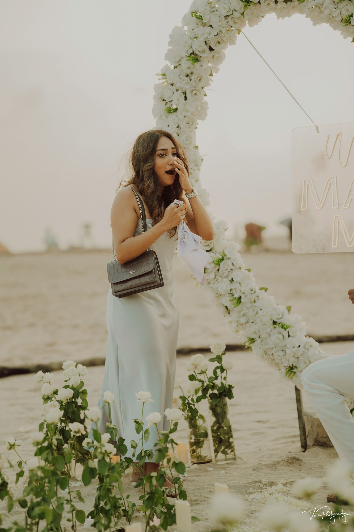 Woman in white dress stands under floral arch on beach with white flowers and greenery in dreamy evening light.
