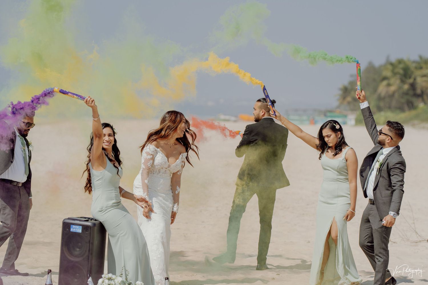 Group of people celebrate on a beach with colorful smoke bombs creating a vibrant and joyful atmosphere at a wedding.