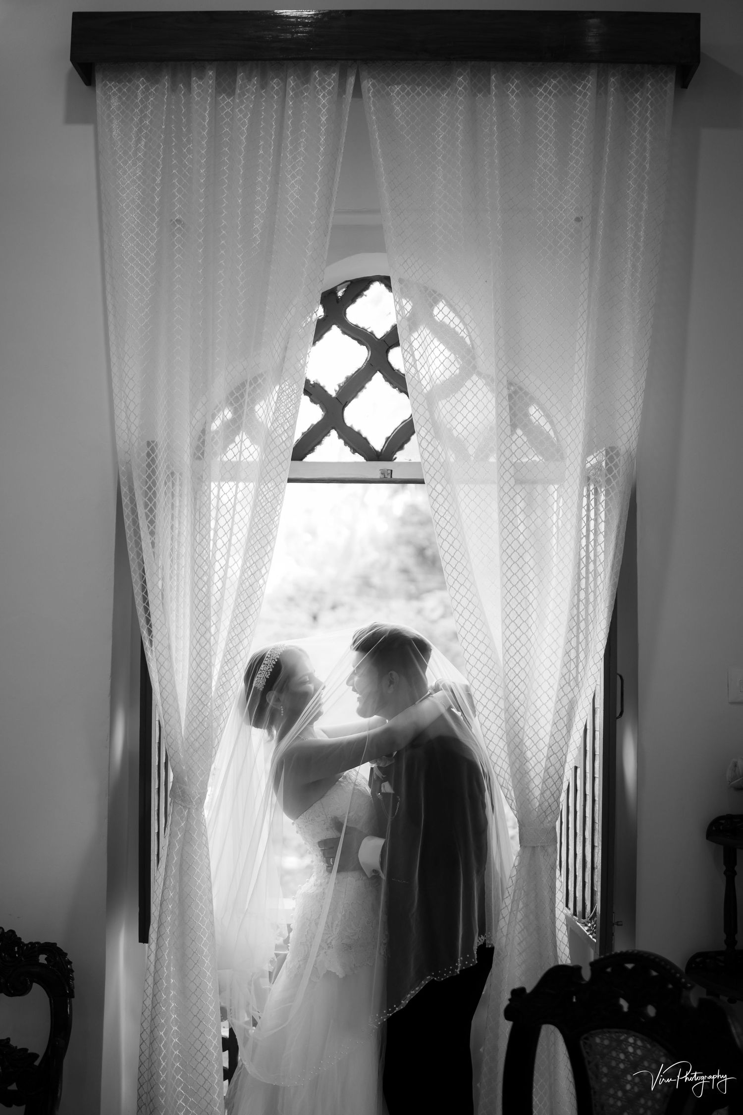 Black and white silhouette of a couple embracing behind sheer curtains in front of a gothic arched window.