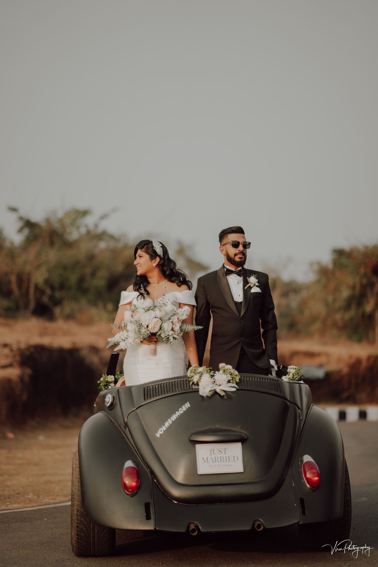 A couple poses in a vintage matte black convertible beetle car at sunset for wedding photos.