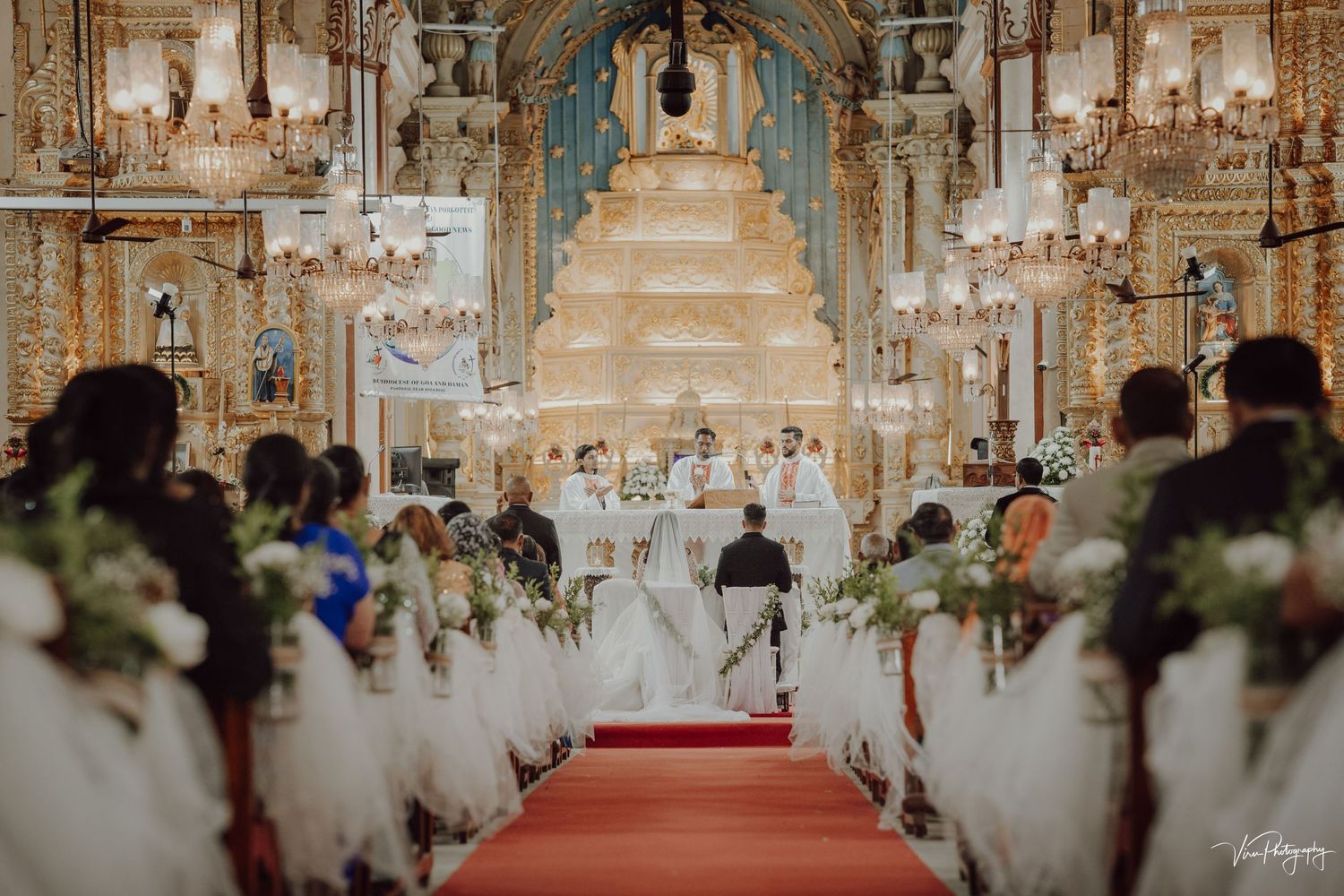 Wedding ceremony in an ornate church with a grand chandelier and white-decorated aisle leading to a tiered altar.