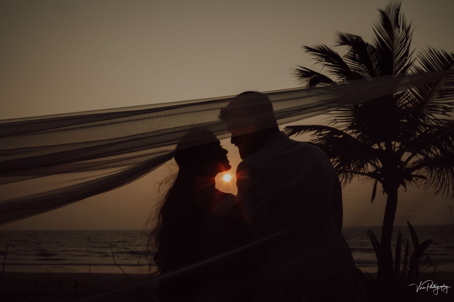 A silhouetted couple shares a romantic moment at sunset on a beach with a palm tree in the background.