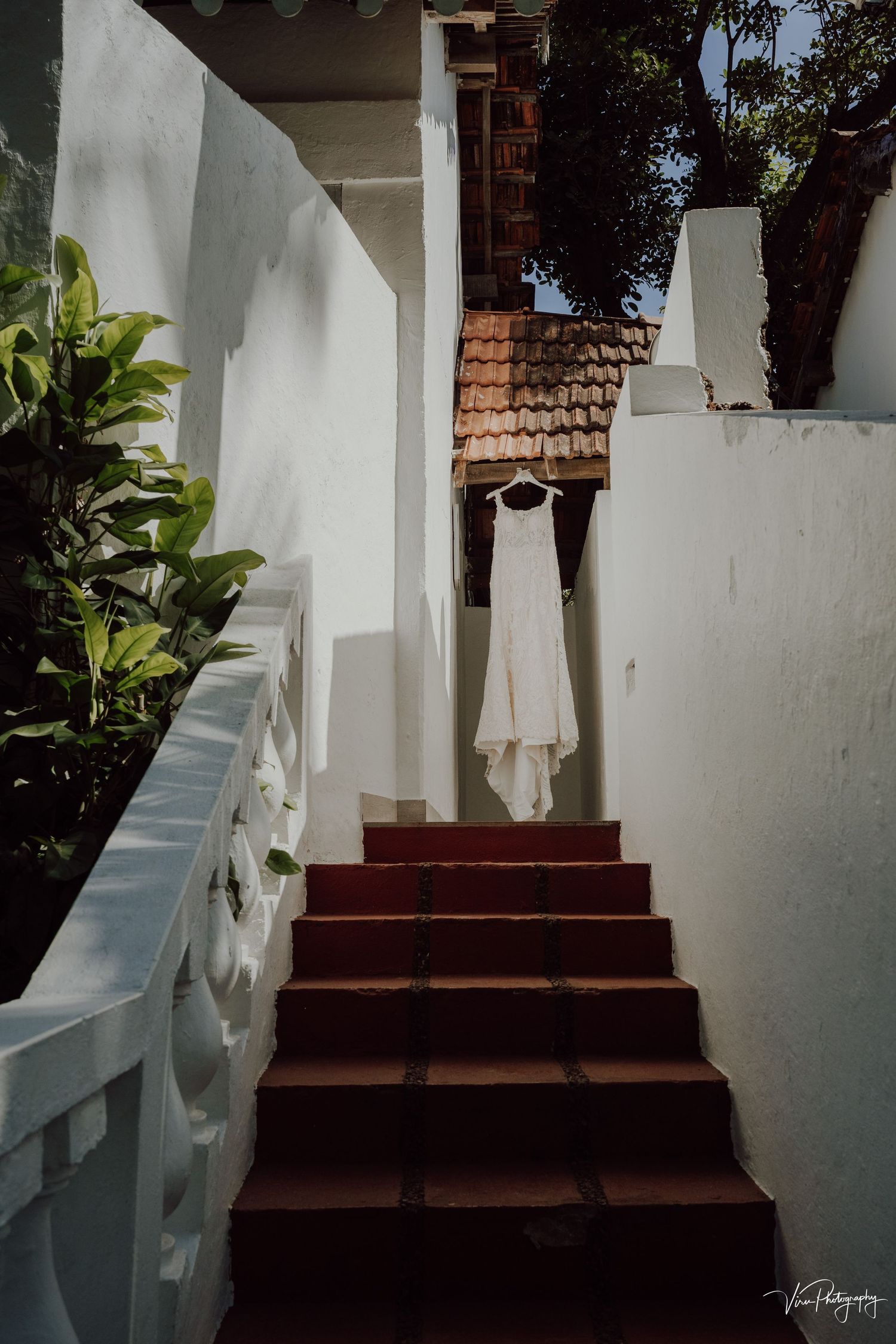 A white wedding dress hangs elegantly at the top of red stairs with white stucco walls and tropical foliage.
