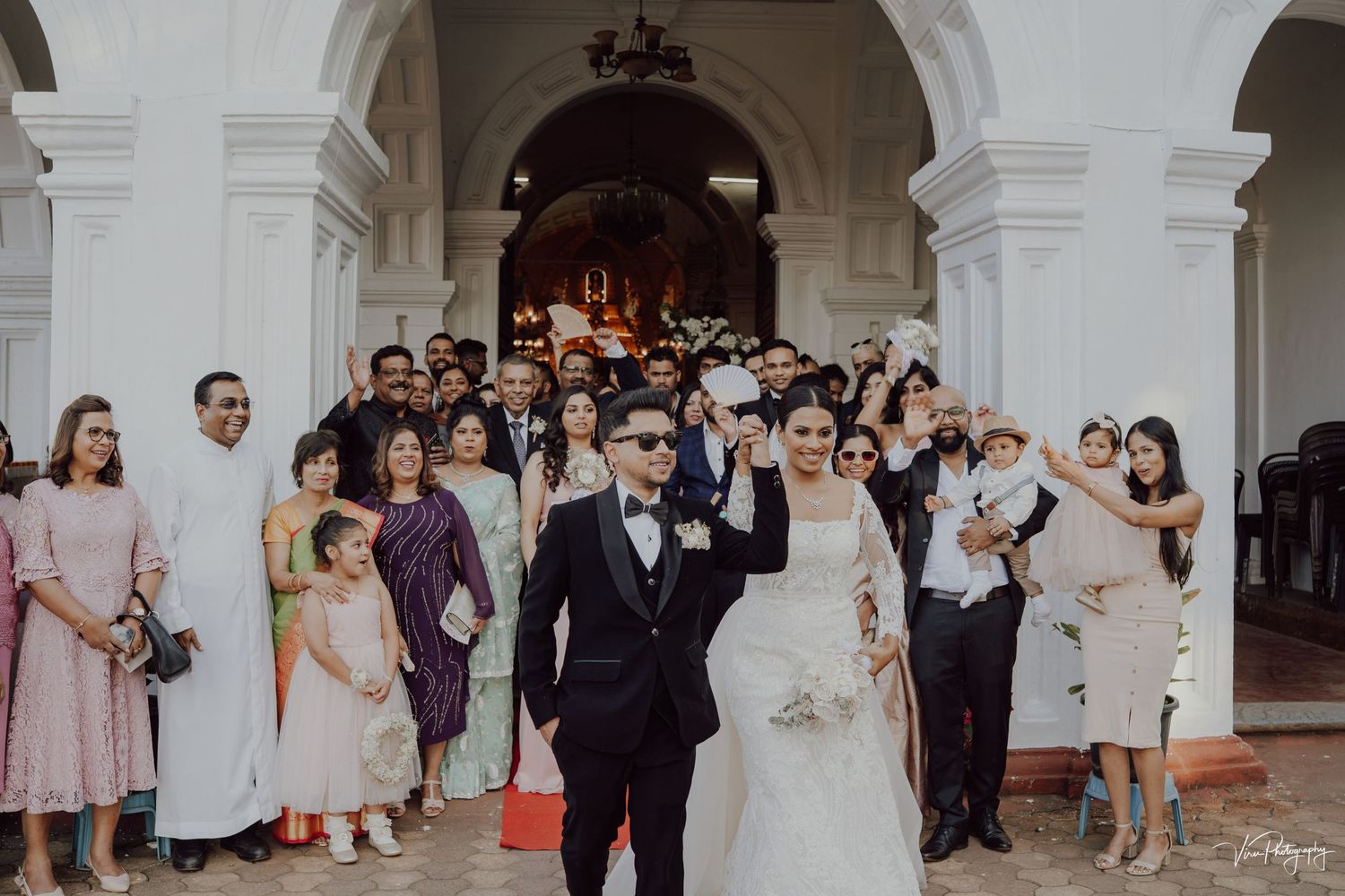 Newlyweds celebrate with their wedding party under elegant white arches at a traditional church ceremony.
