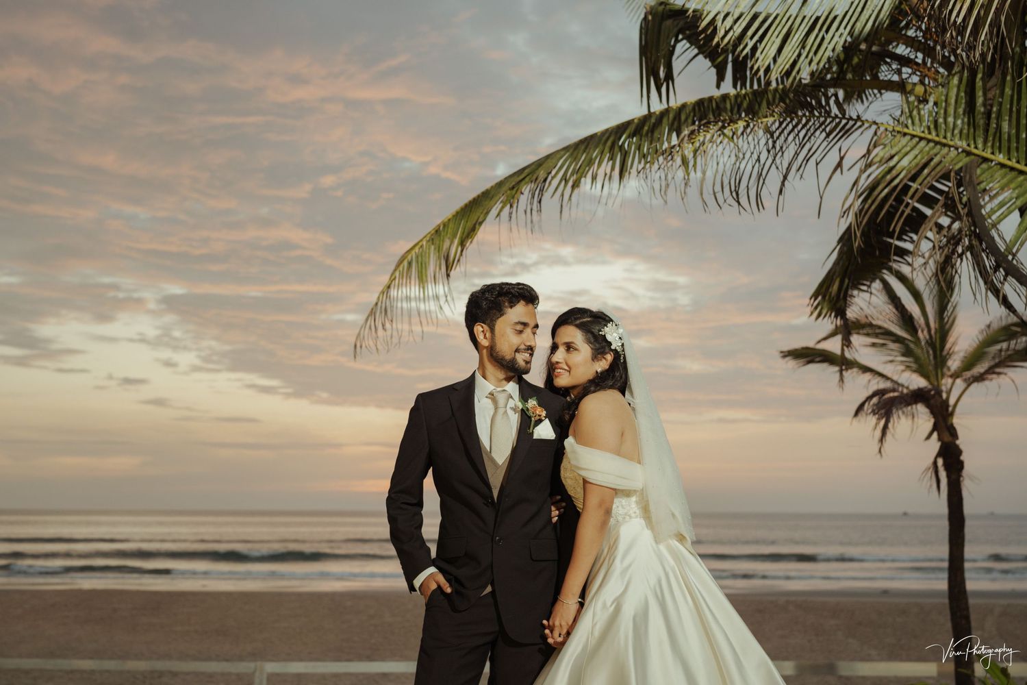 A romantic beach wedding photo with palm trees silhouetted against a warm sunset sky at the ocean shore.