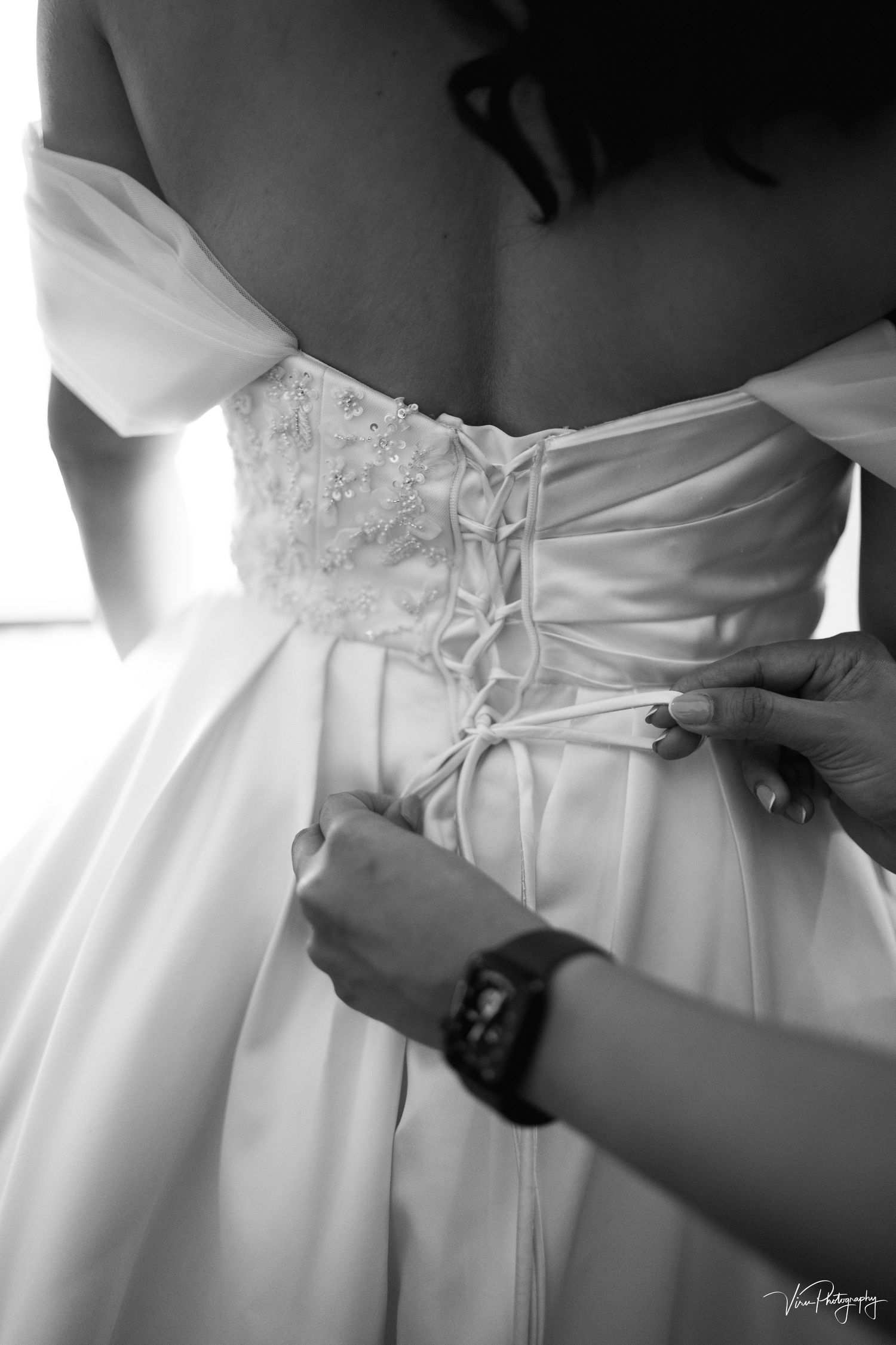 A black and white photograph shows hands lacing up the corset back of an elegant wedding dress.