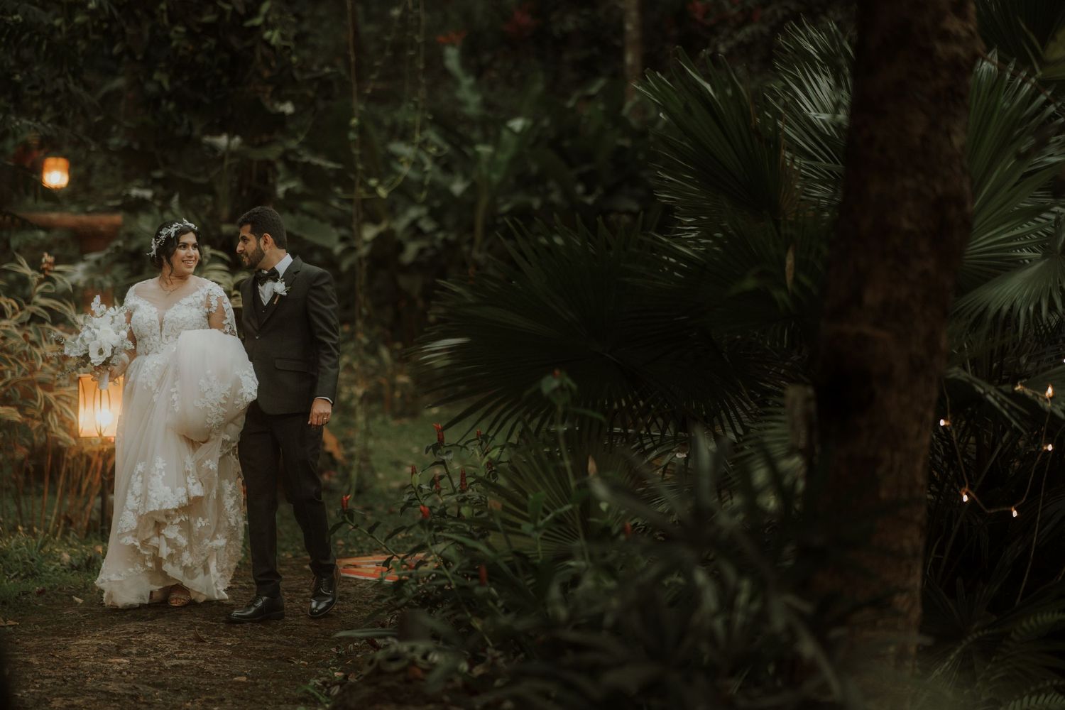 Newlyweds walk through a tropical garden setting lit by hanging lanterns at dusk.