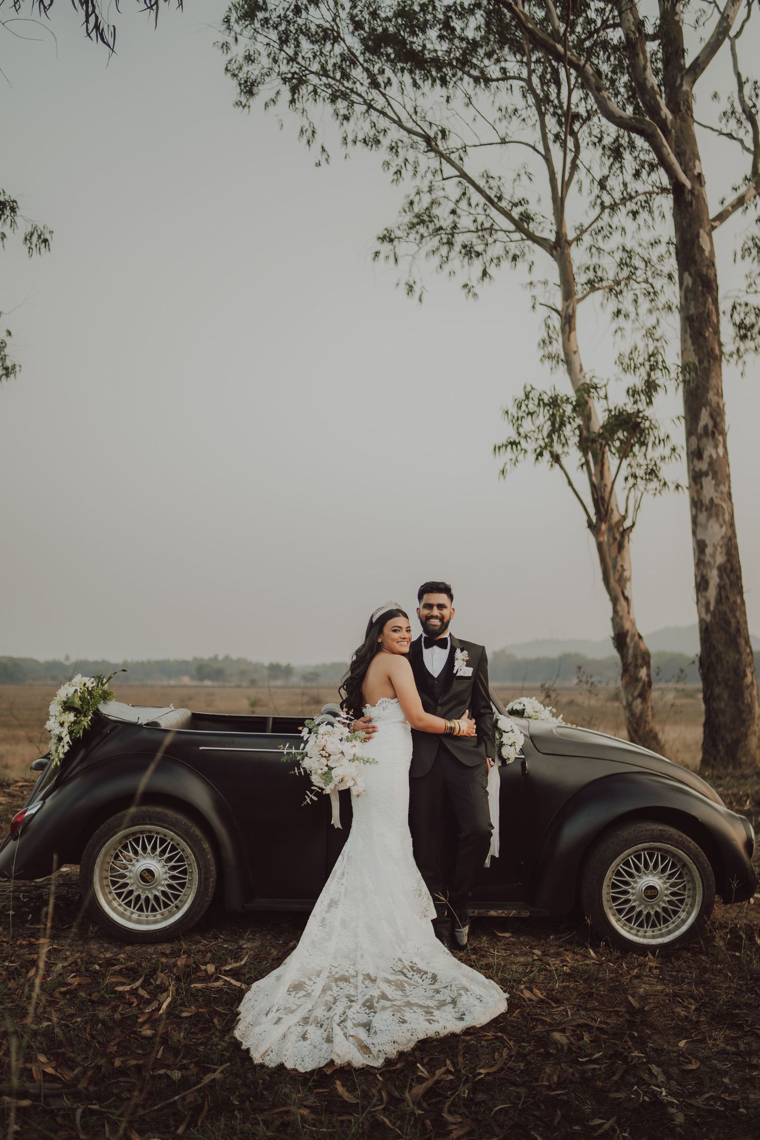 A wedding couple poses with a vintage black car against a backdrop of eucalyptus trees.