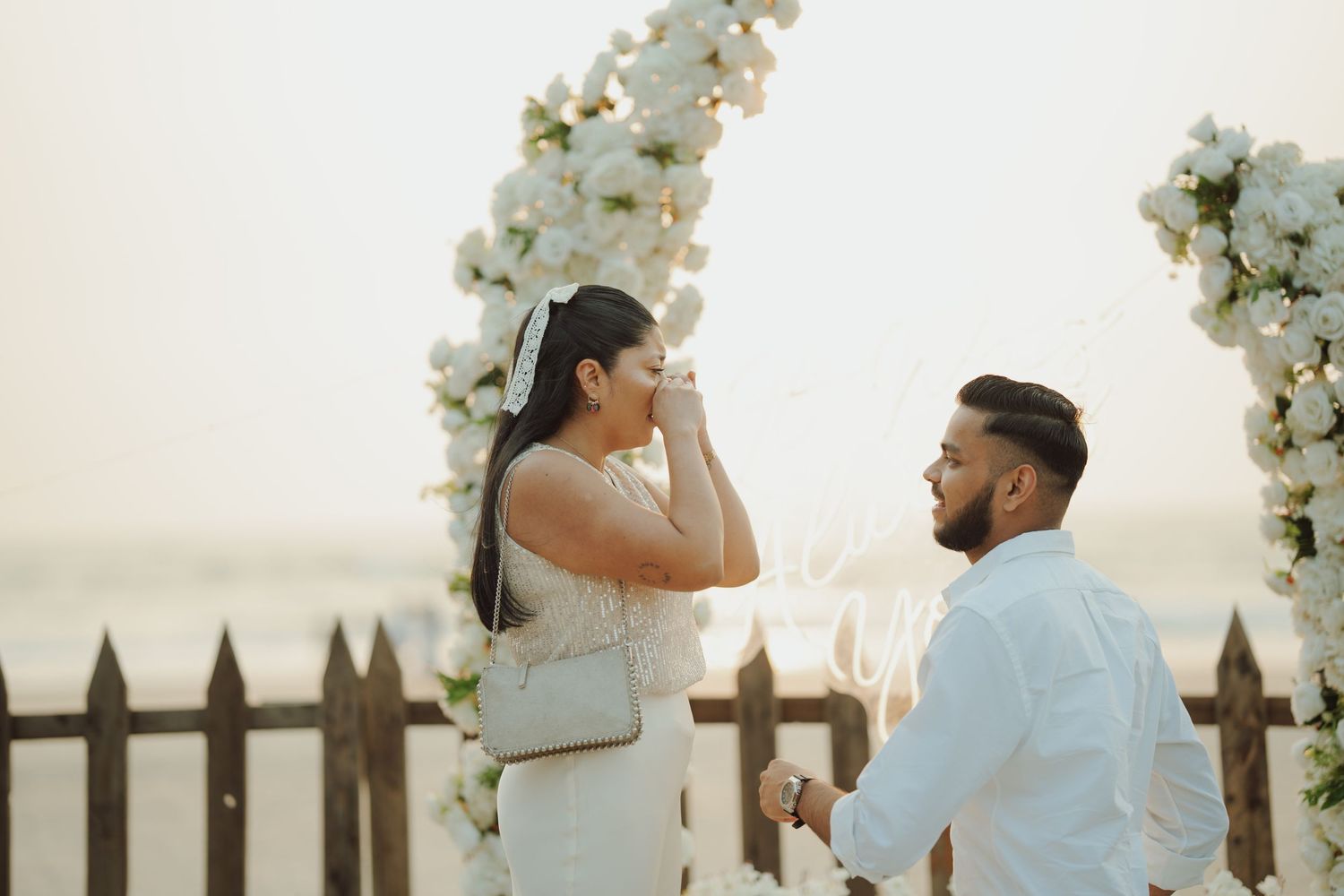 Man proposes to emotional woman on beach near white floral arch at sunset.