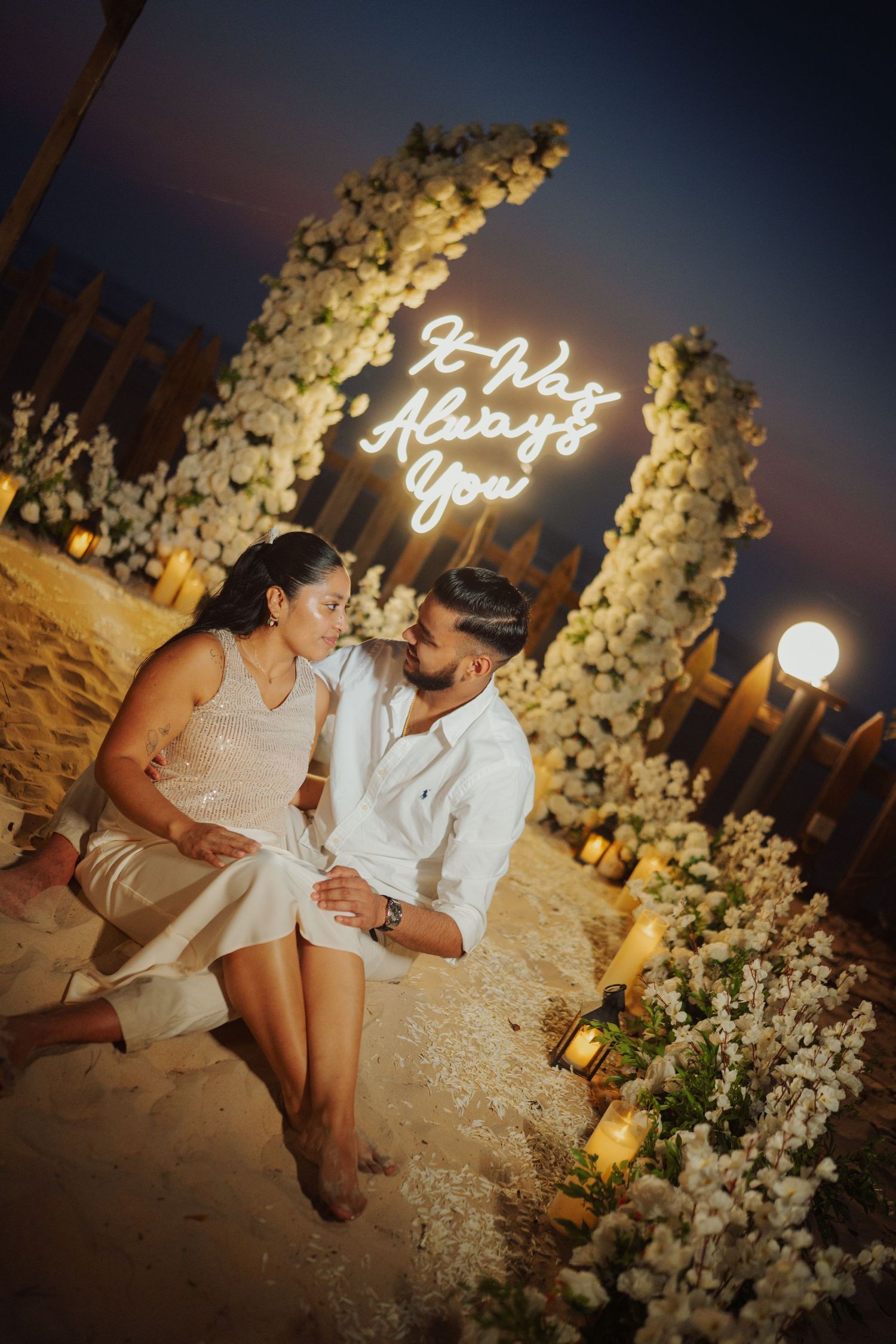 Romantic couple sitting together at night beside a floral arch with neon sign reading It Was Always You on a beach.
