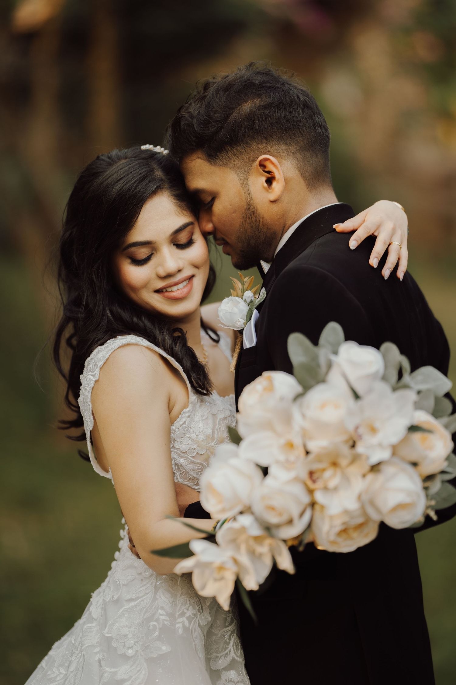 A romantic wedding couple embraces while holding a bouquet of pale peach flowers.