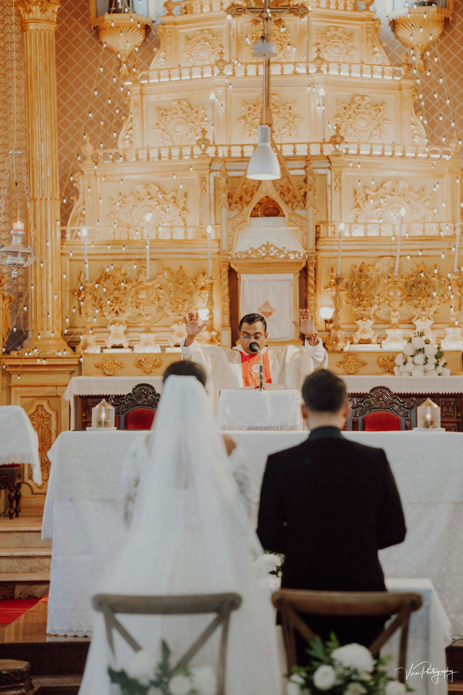 Catholic wedding ceremony taking place in an ornate golden church altar with attendees seated before the priest.