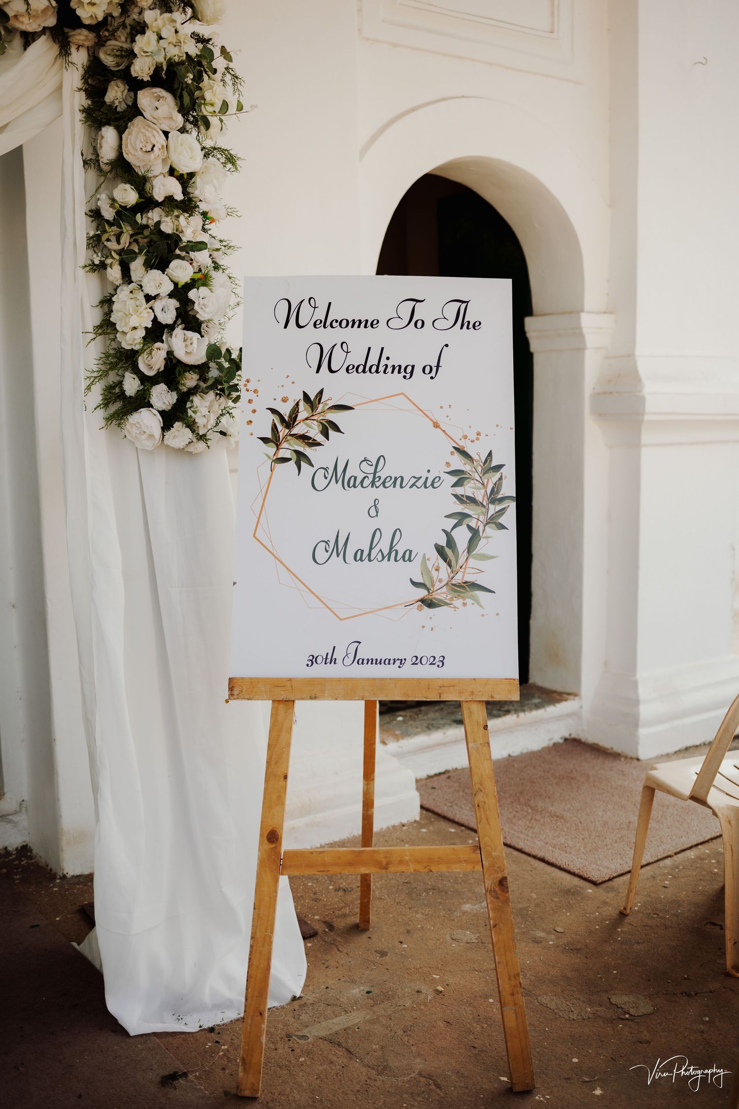 Elegant wedding welcome sign on wooden easel with white florals and greenery arch decoration against white wall.