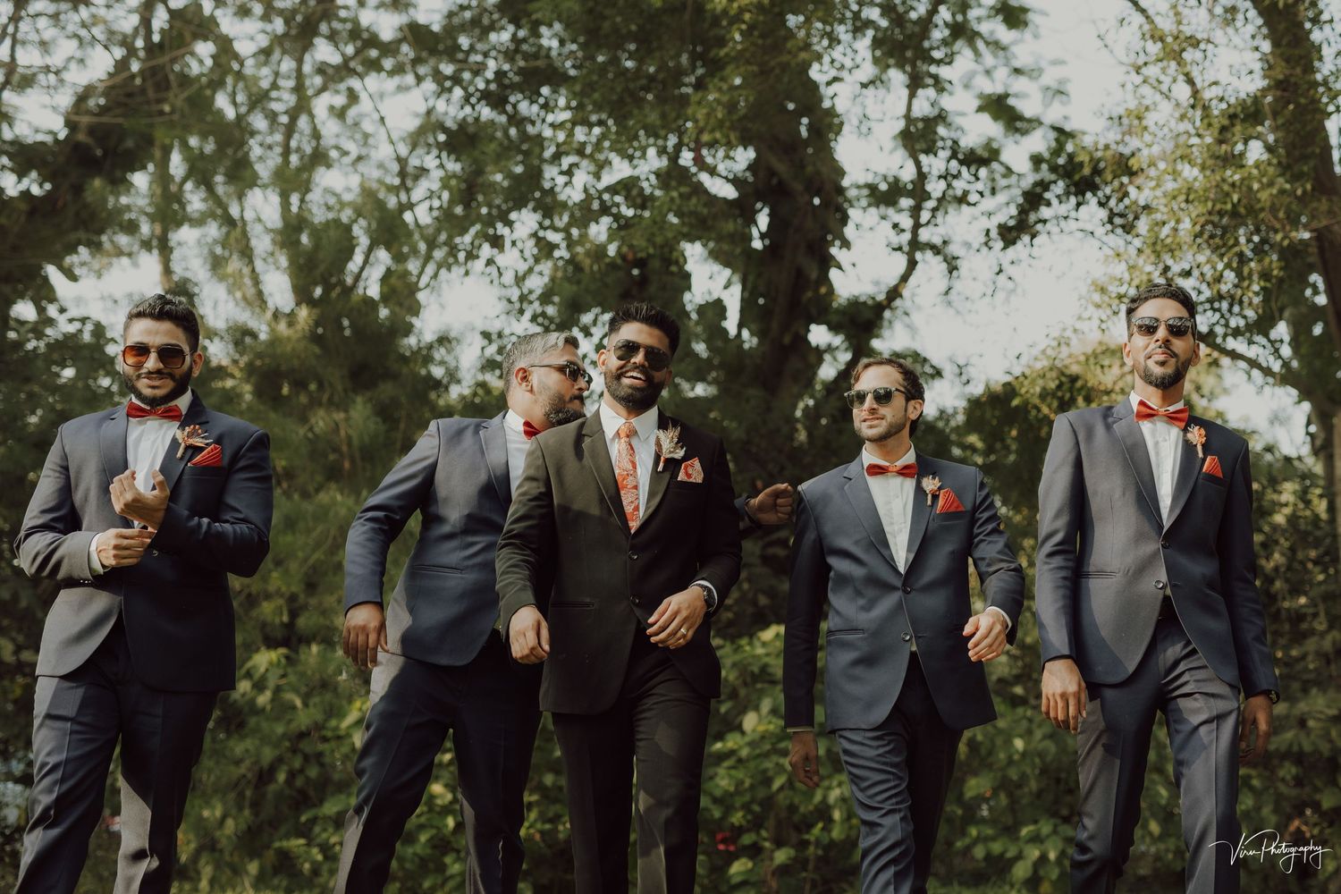 Five groomsmen in fitted suits walking together on a tree-lined path during a sunny outdoor wedding celebration.