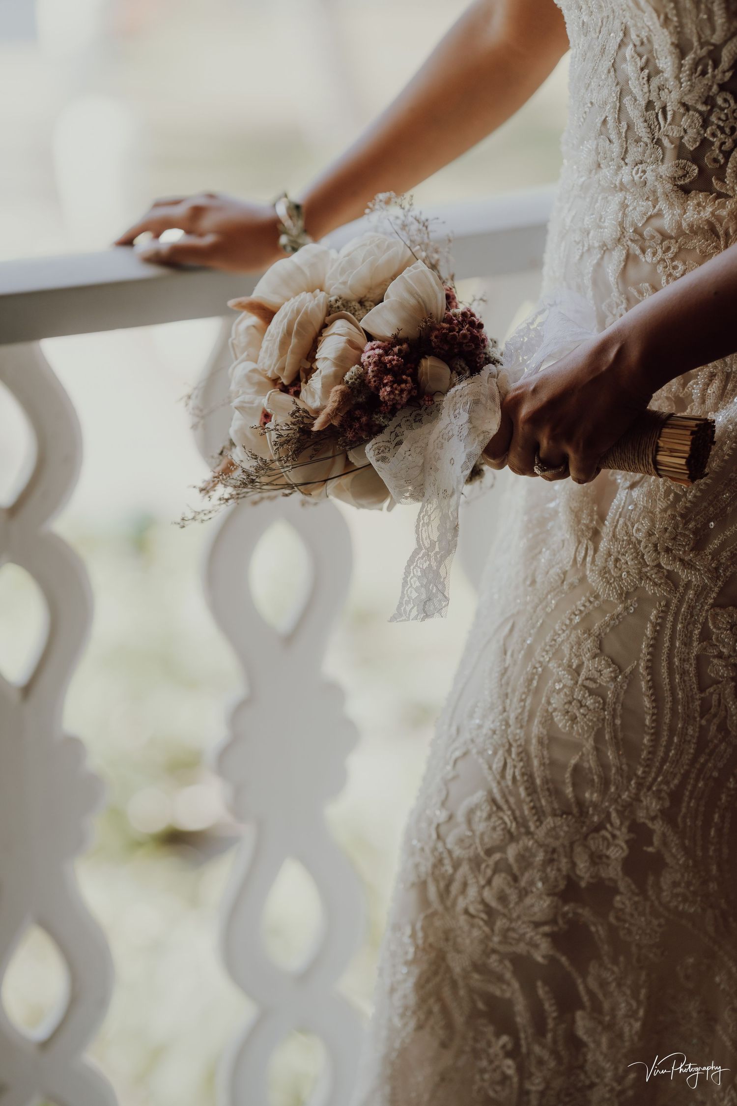 Bride in lace wedding dress holds romantic rustic bouquet of cream roses and burgundy flowers on white balcony.