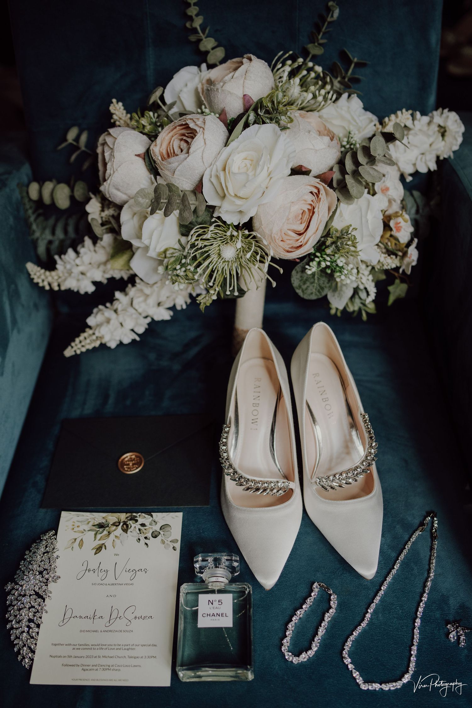 Elegant wedding details including white rose bouquet, pearl-adorned heels, perfume bottle, and invitation on dark blue fabric.