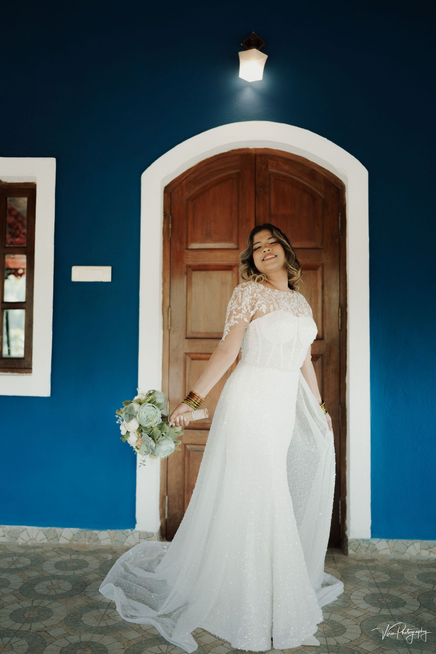 A bride in an elegant white lace wedding gown poses against a vibrant blue wall and wooden doorway with a bouquet.