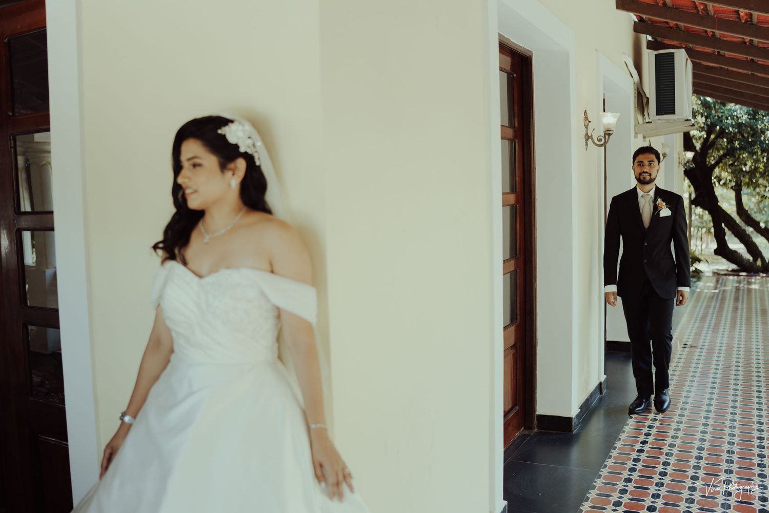 A dramatic wedding scene with a white gown against a white wall, with a suited figure visible in the background.