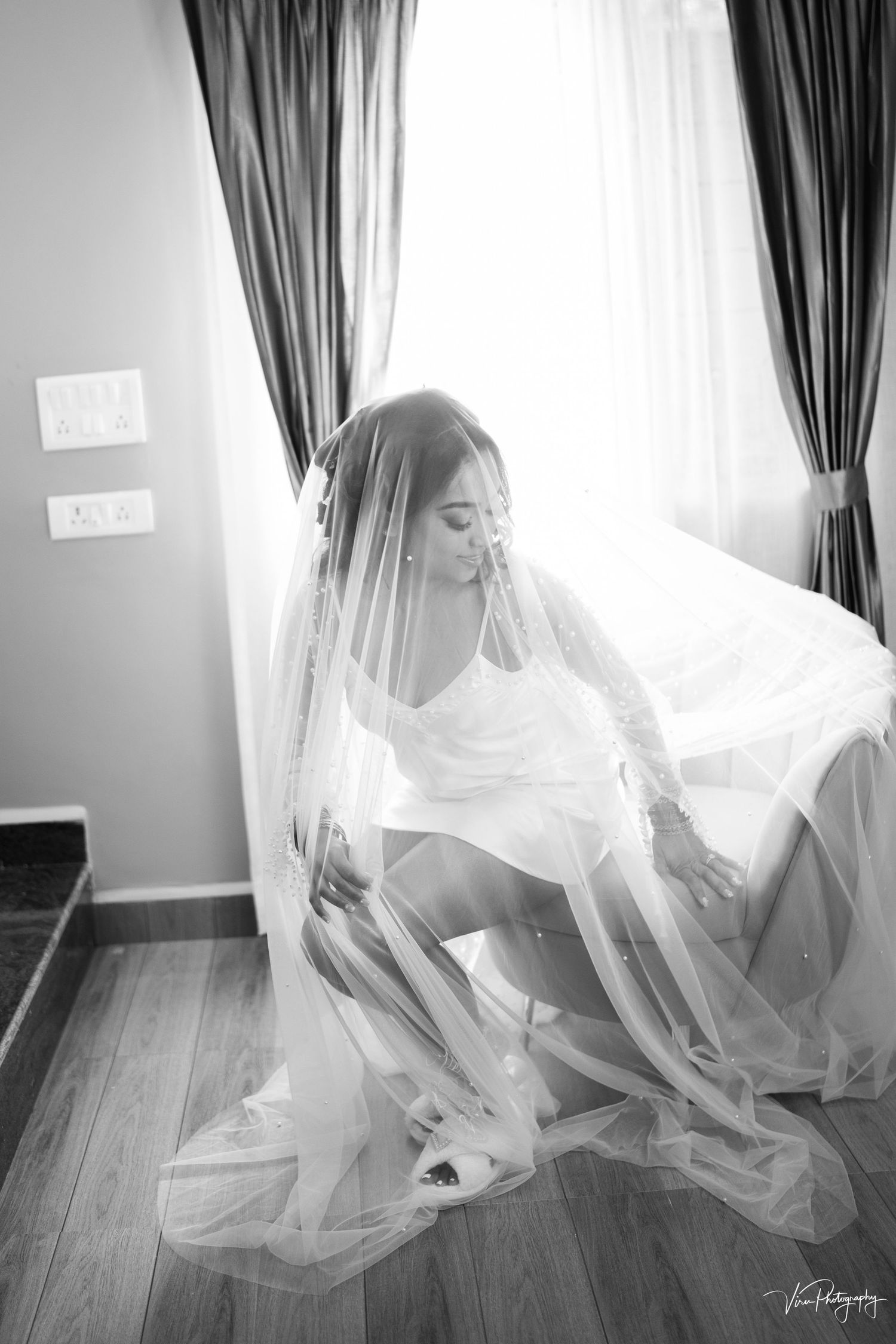 Ethereal bride in flowing white veil kneels by window light in black and white photography.