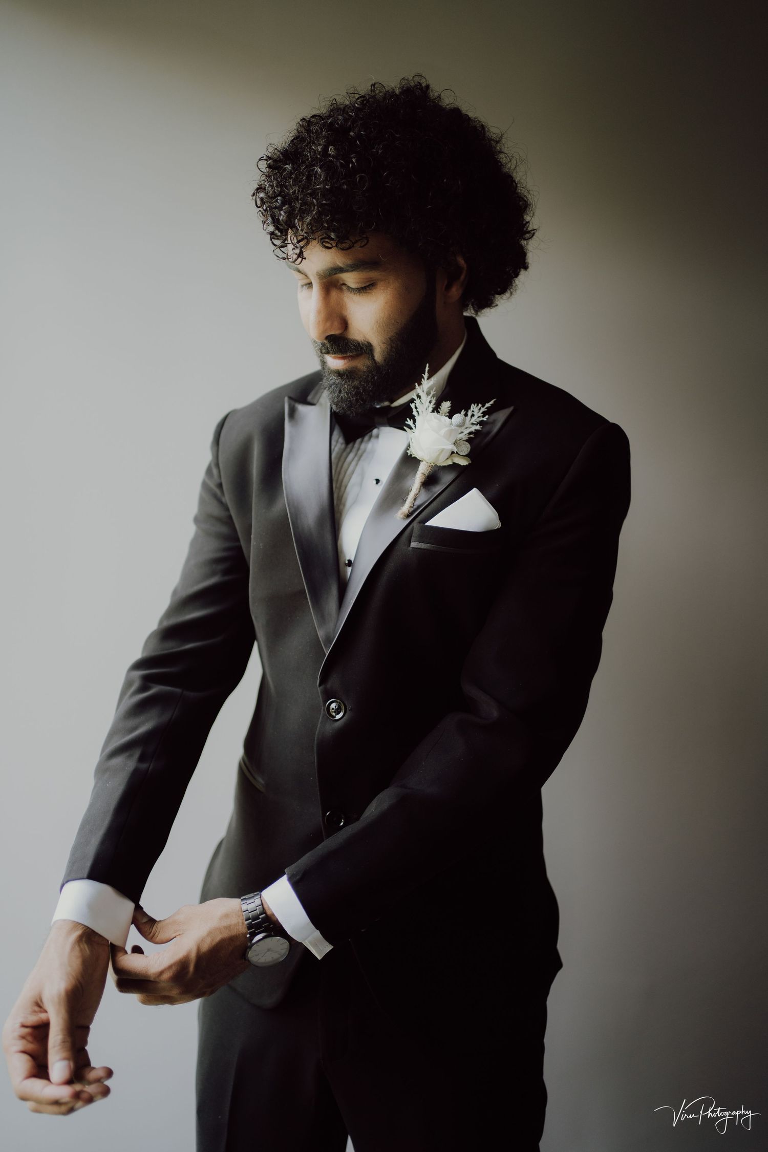 A groom in a classic black tuxedo with a white boutonniere adjusts his cuff links in a moody portrait.