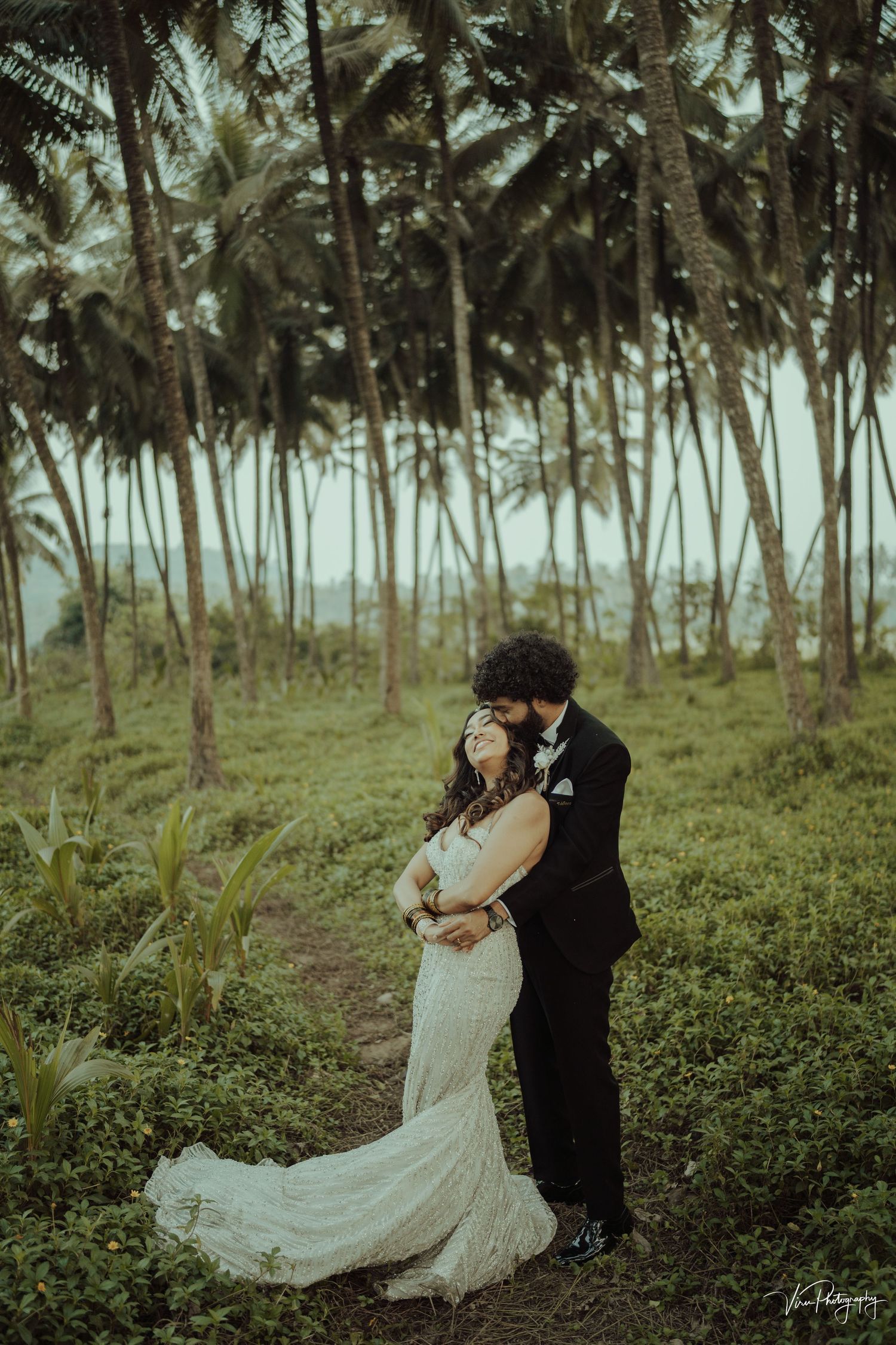 A romantic couple embraces in a tropical palm grove, with a lace wedding dress trailing on the lush green grass.
