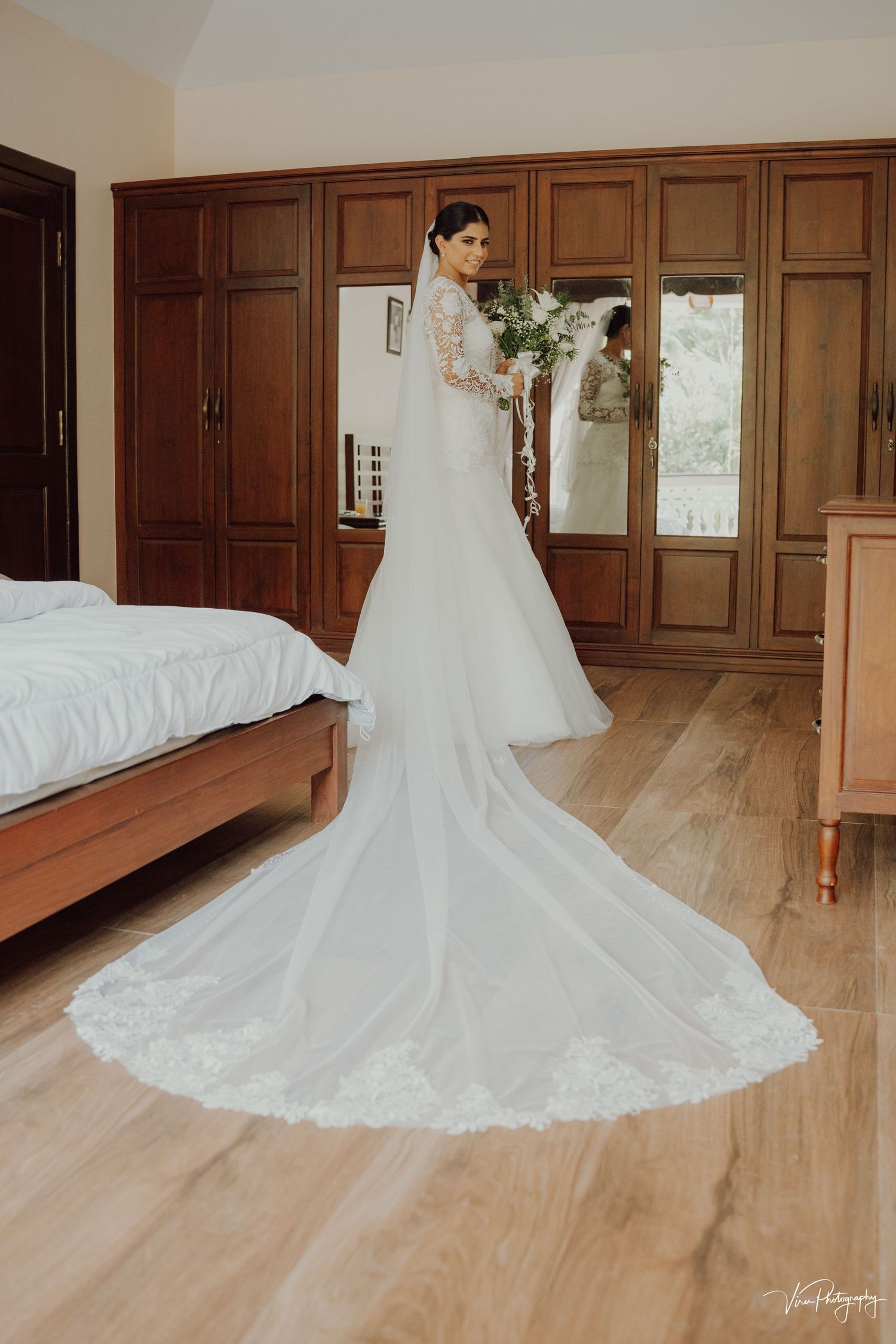 Elegant white wedding gown with long lace-trimmed train on hardwood floor in bedroom with wooden paneled walls.