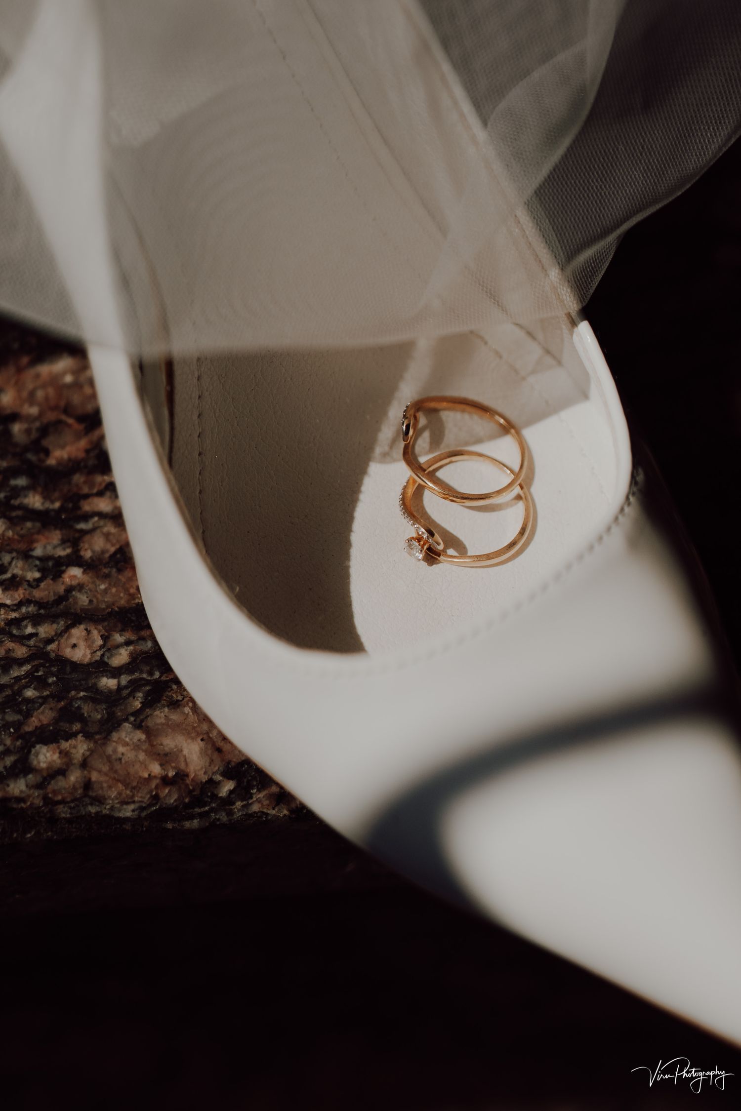 Gold wedding rings elegantly placed on a white bridal shoe against a dark textured background with soft veil details.