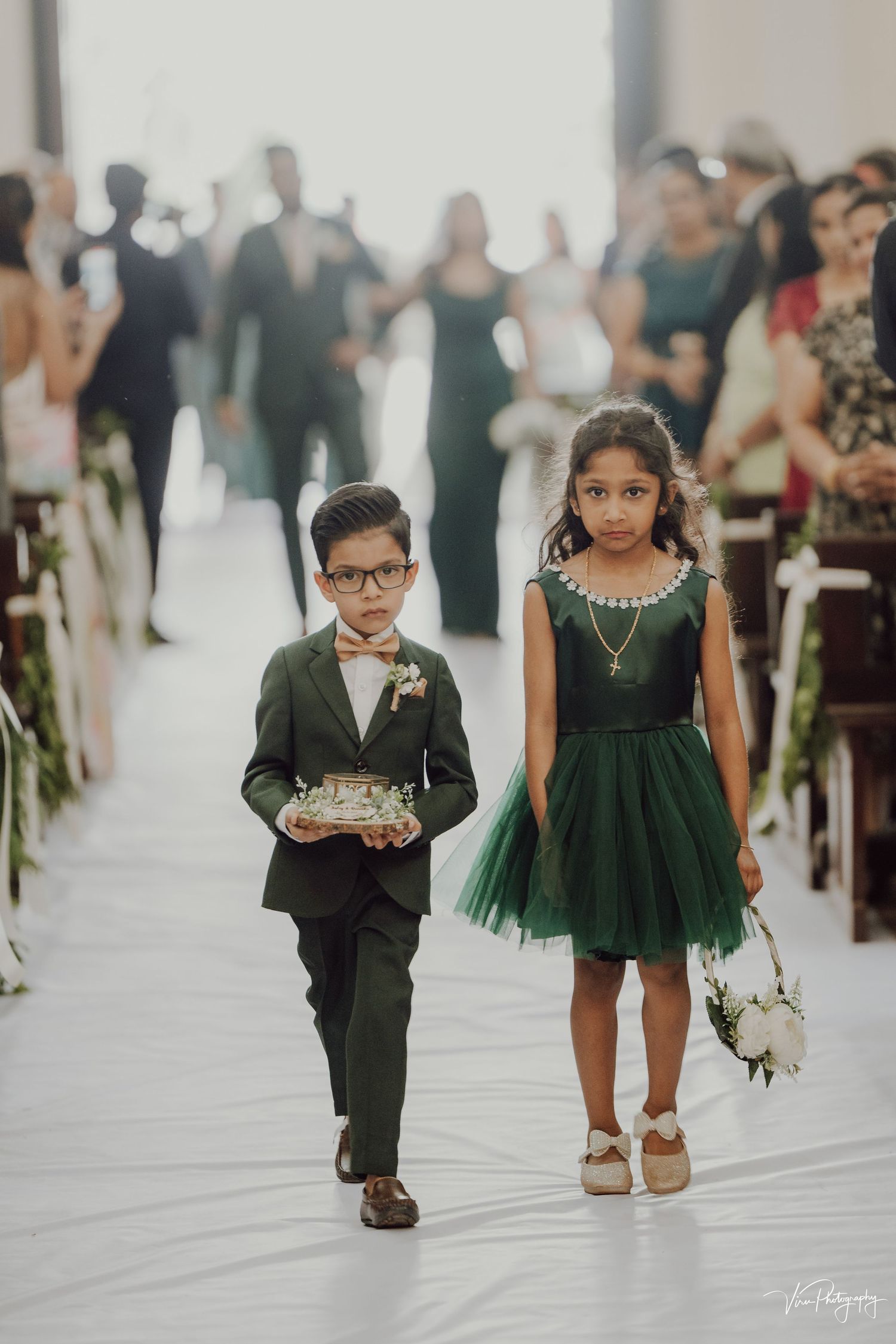 Ring bearer in suit and flower girl in green dress walking down white aisle at formal wedding ceremony.
