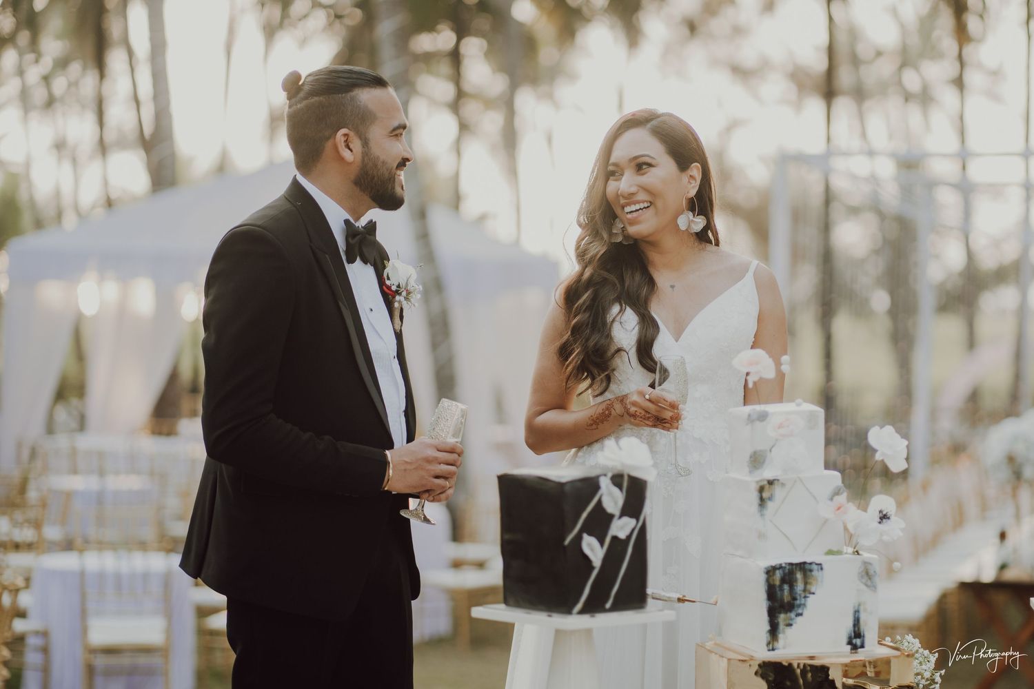 A bride and groom share a laugh together next to their white wedding cake at an elegant outdoor reception.