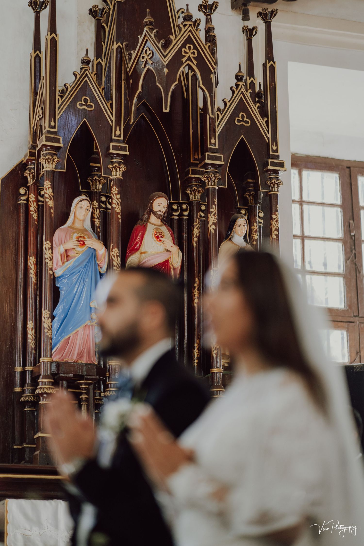 Ornate Gothic church altar with religious statues in niches while couple participates in wedding ceremony in foreground.