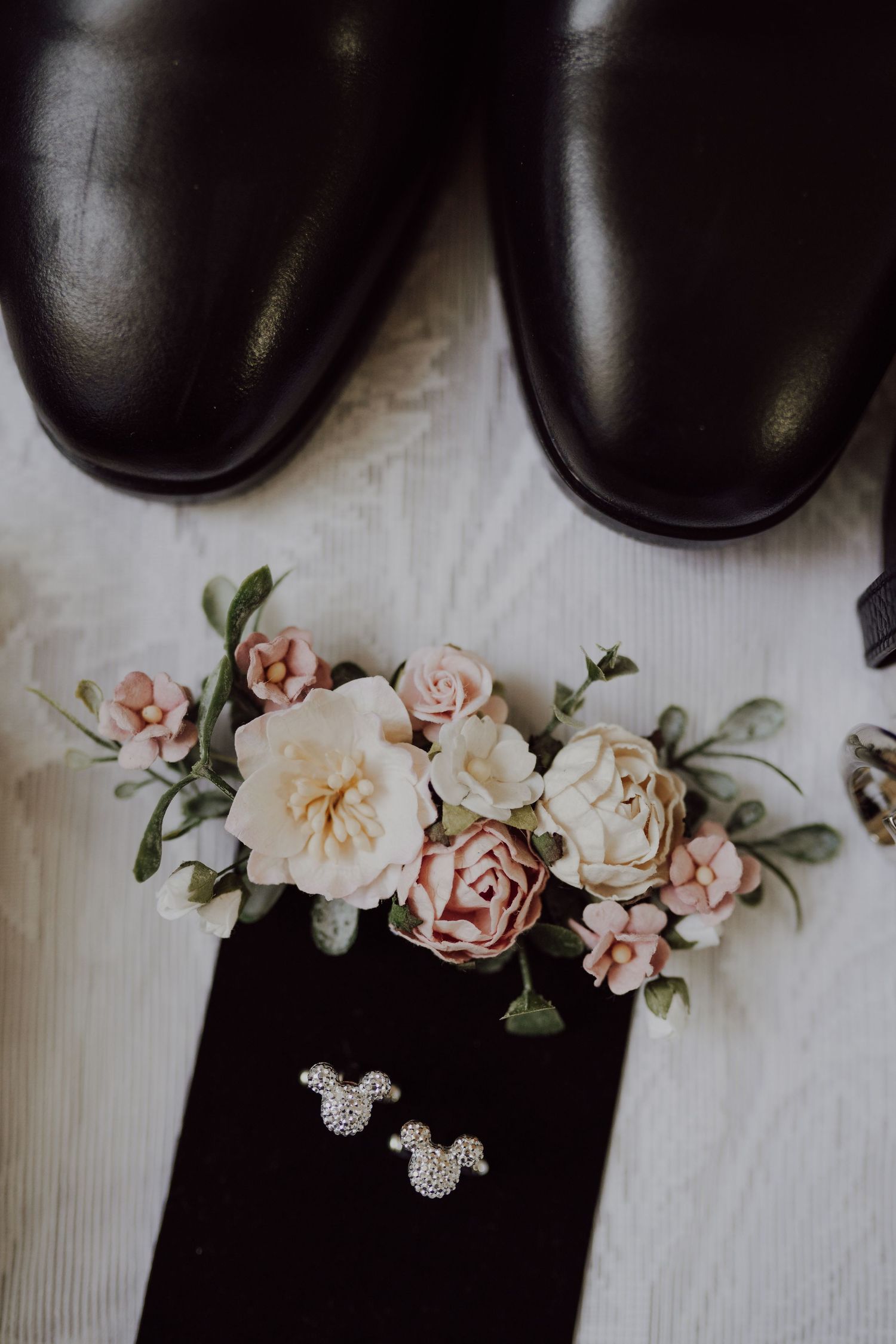 Black dress shoes with delicate pink and white flowers and Mickey Mouse shaped silver earrings arranged on white fabric.