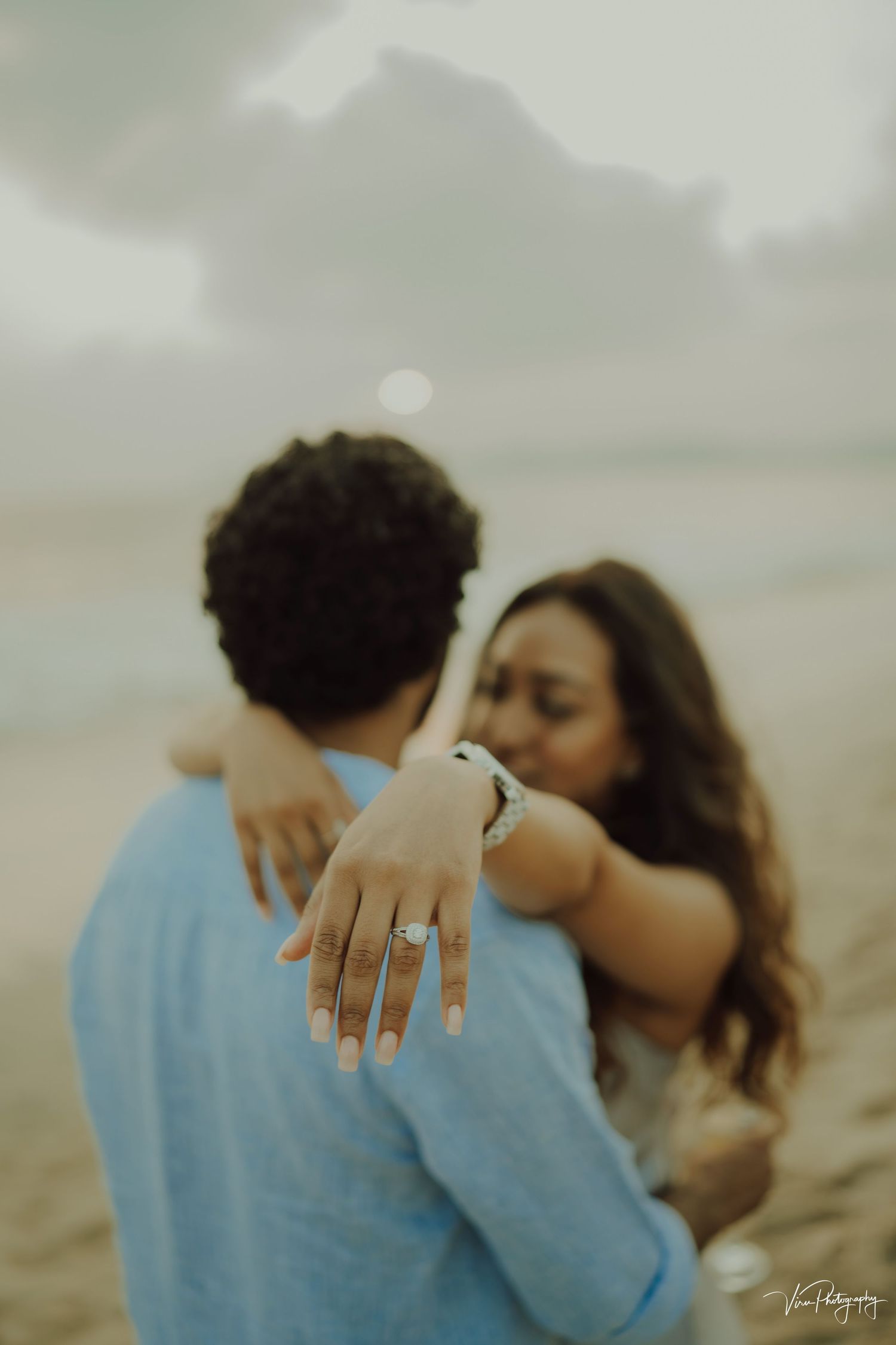 Engaged couple embraces on misty beach as woman shows off ring during romantic proposal photoshoot.