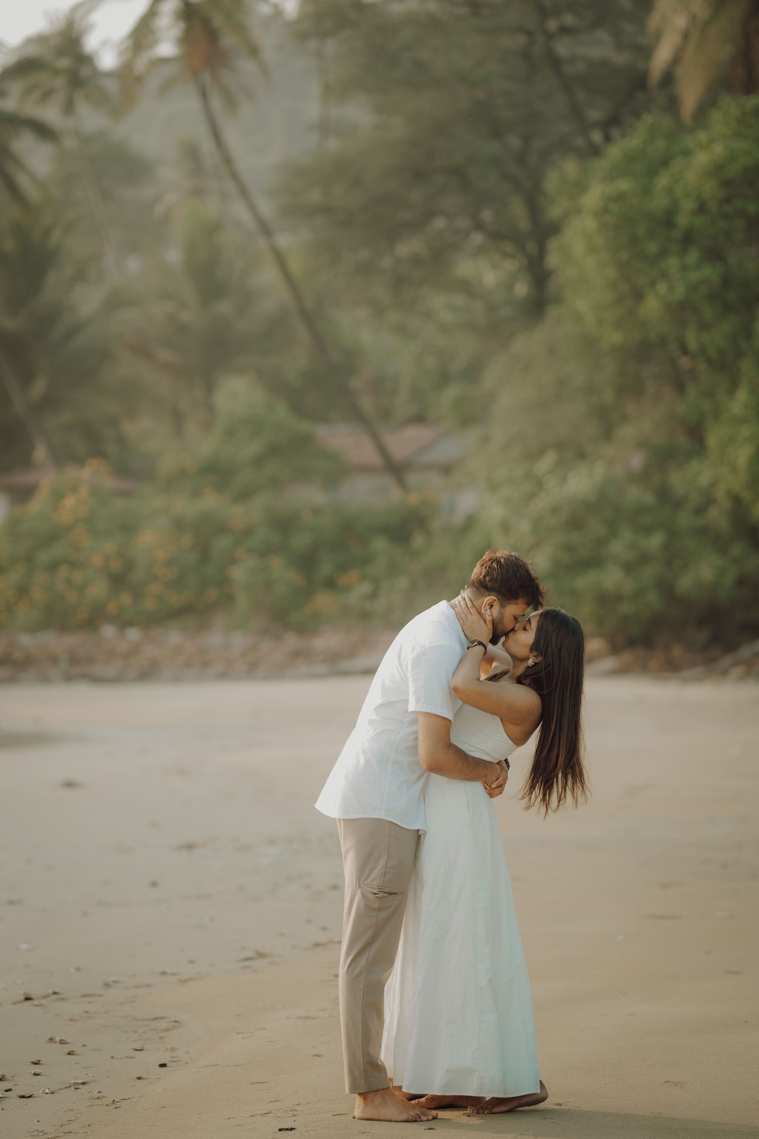A romantic couple shares an intimate moment on a misty tropical beach in white and beige attire.