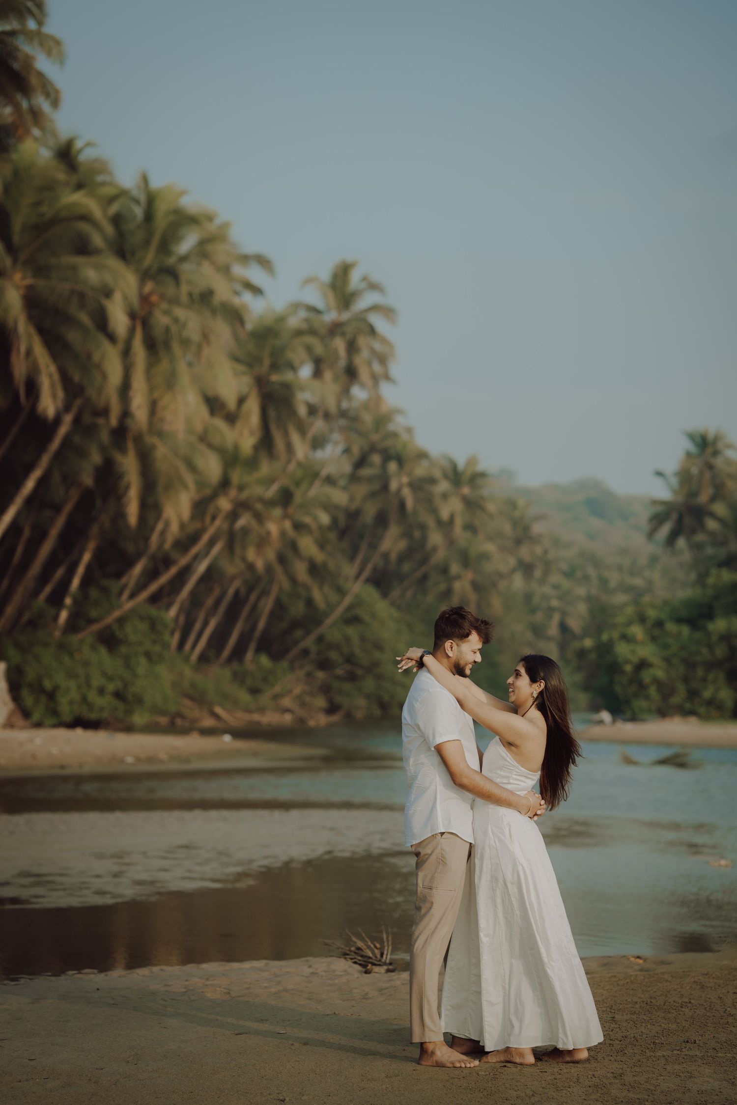 A couple in white outfits shares a romantic moment on a tropical beach with palm trees and mountains in the background.